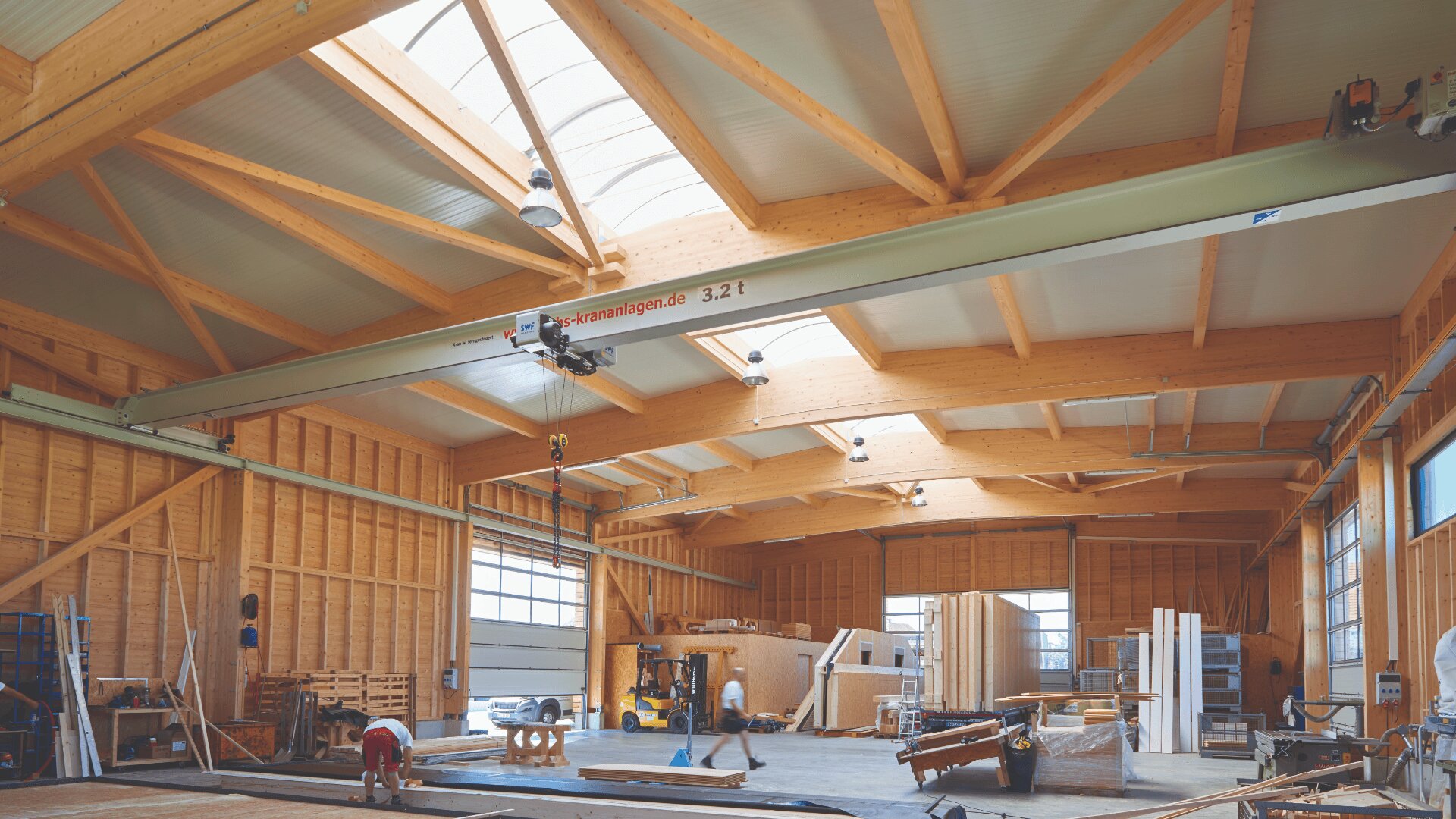 Eine geräumige, gut beleuchtete Holzwerkstatt mit hohen Holzdecken, großen Fenstern, einem Deckenkran und Arbeitern, die mit Holz und Geräten arbeiten. Überall in der Werkstatt sind Holzstapel und verschiedene Werkzeuge zu sehen.