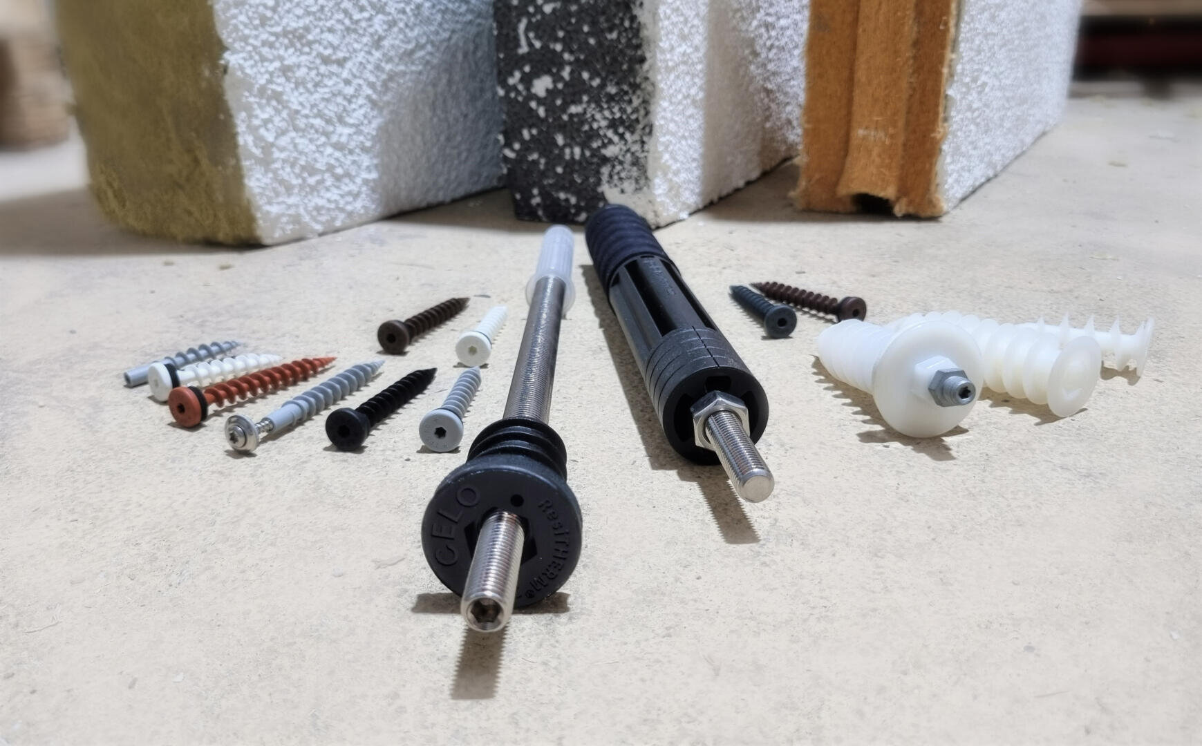 Auf einer Fläche mit drei Baustoffmustern im Hintergrund, darunter Beton, Ziegel und Dämmplatten, sind verschiedene Dübel, Schrauben und Anker ausgestellt.