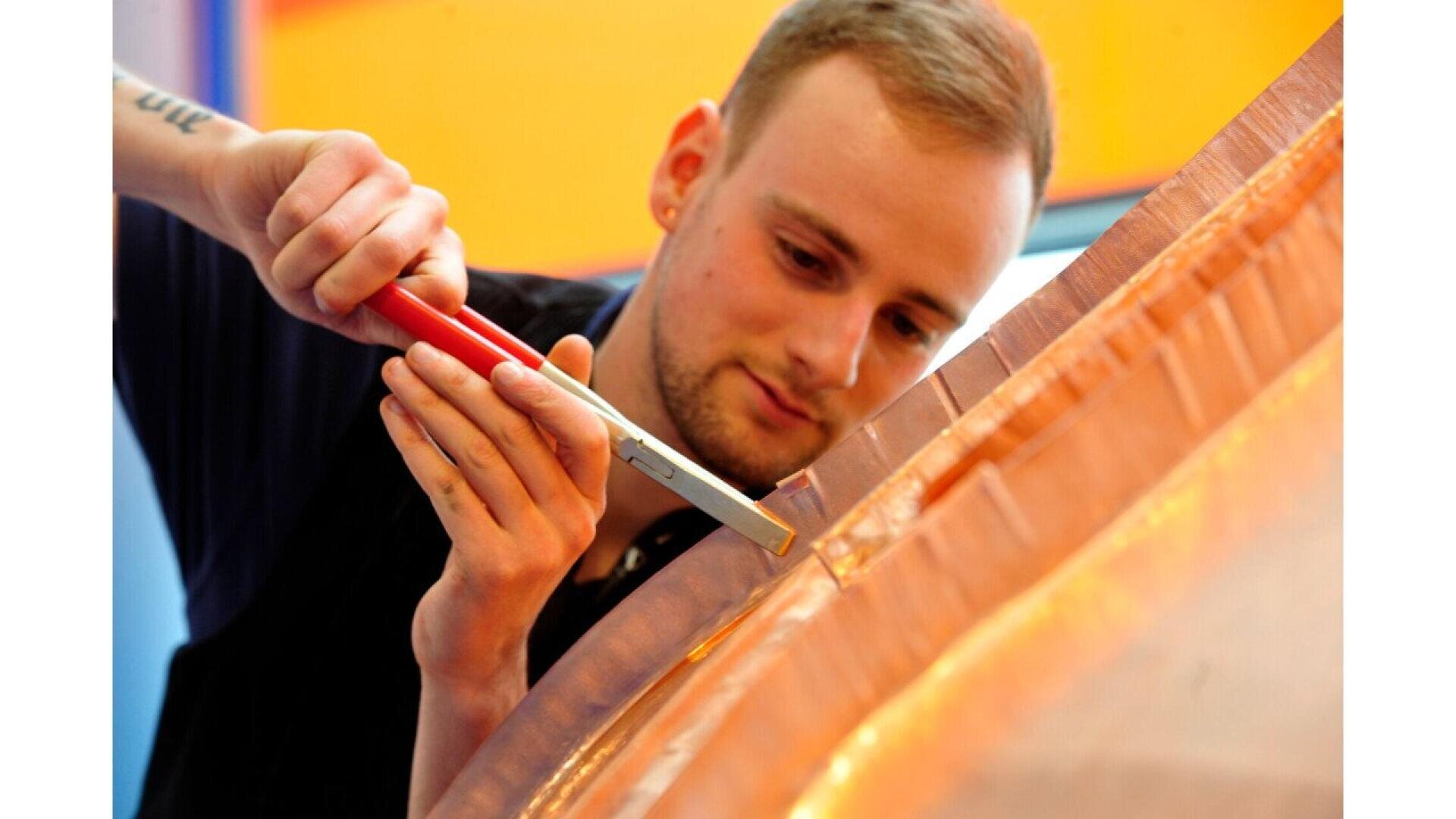 A man uses a red-handled tool to carefully work on a shiny copper surface, focusing intently on his detailed task.
