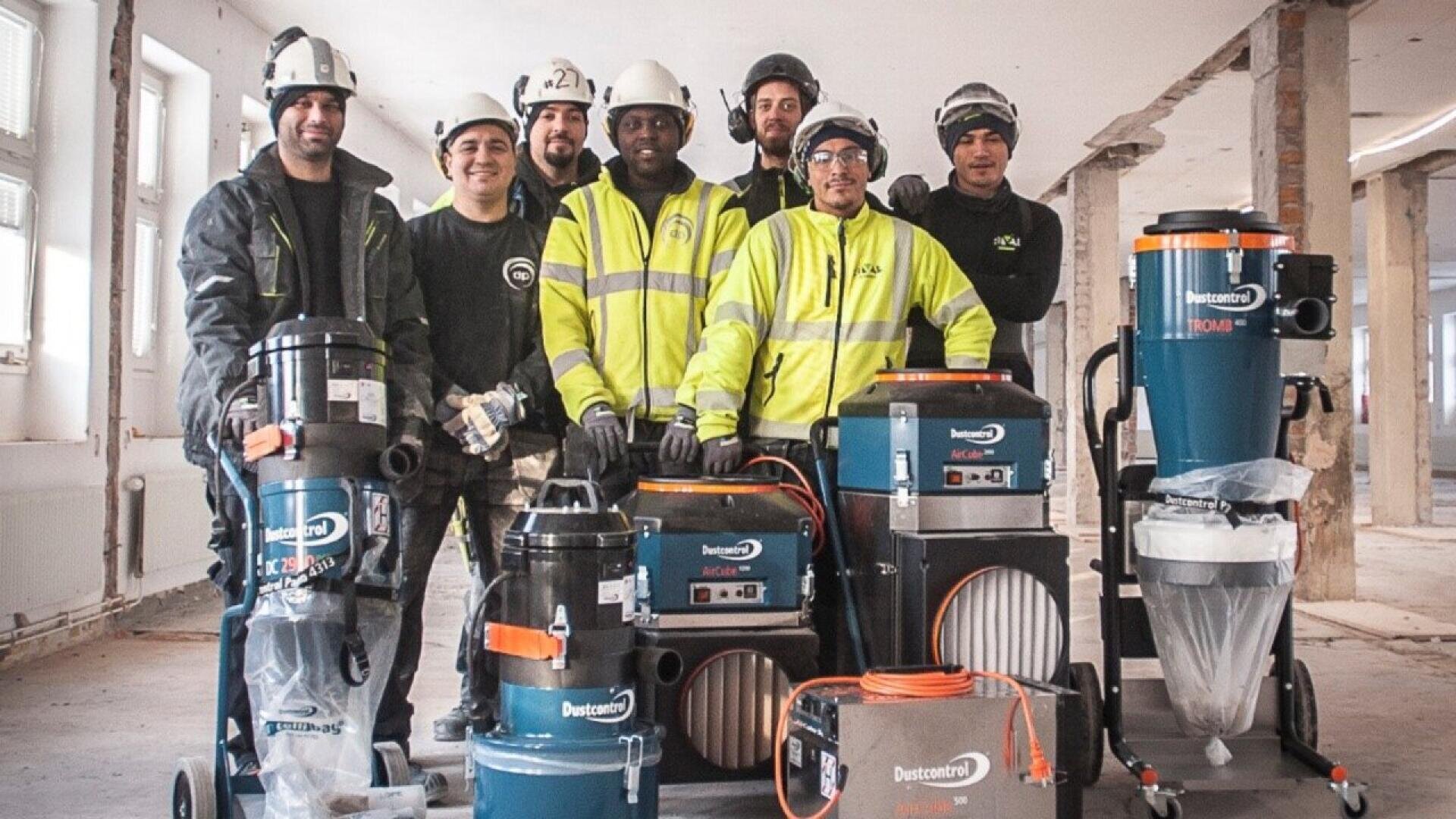A group of seven construction workers in safety gear pose indoors with industrial vacuum equipment and tools in a partially renovated building.