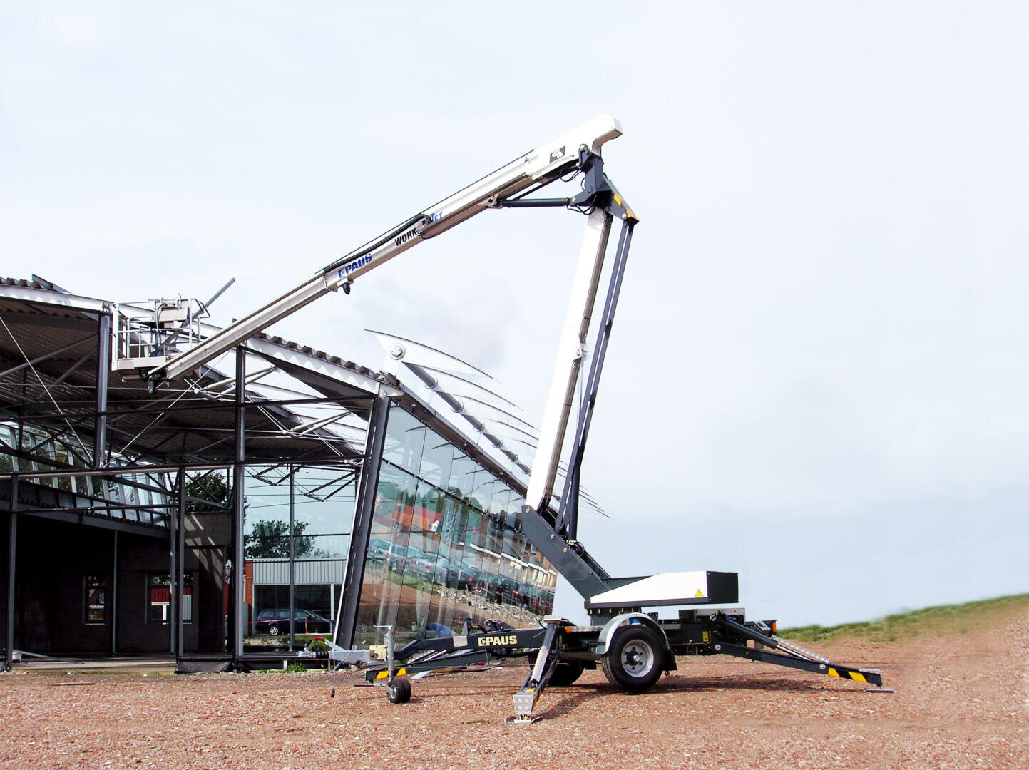 Eine auf einem Anhänger montierte hydraulische Hebebühne steht vor einem modernen Glasgebäude und reicht mit ihrem ausgefahrenen Arm für Wartungs- oder Reparaturarbeiten bis zum Dach.