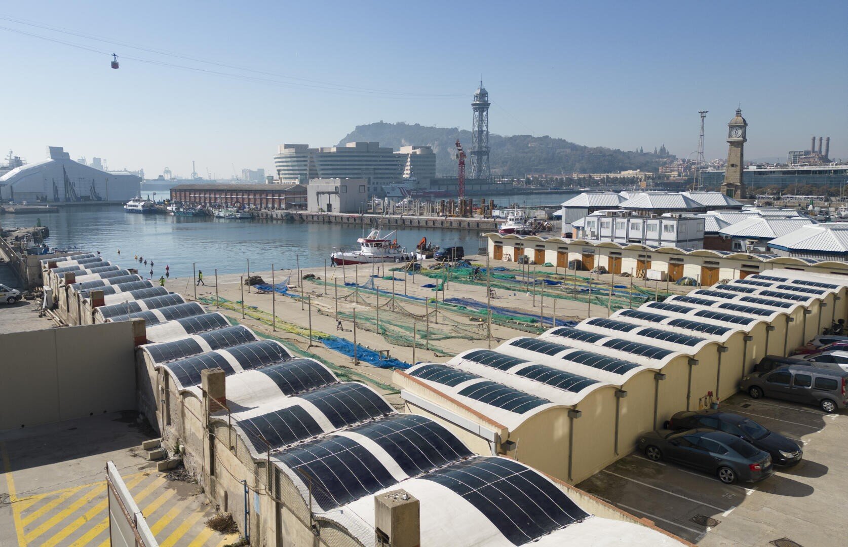 Ein Uferbereich mit Reihen von Gebäuden mit Solarmodulen auf den geschwungenen Dächern, mehreren geparkten Autos, einem Hafen mit Booten und Stadtgebäuden im Hintergrund unter einem klaren Himmel.