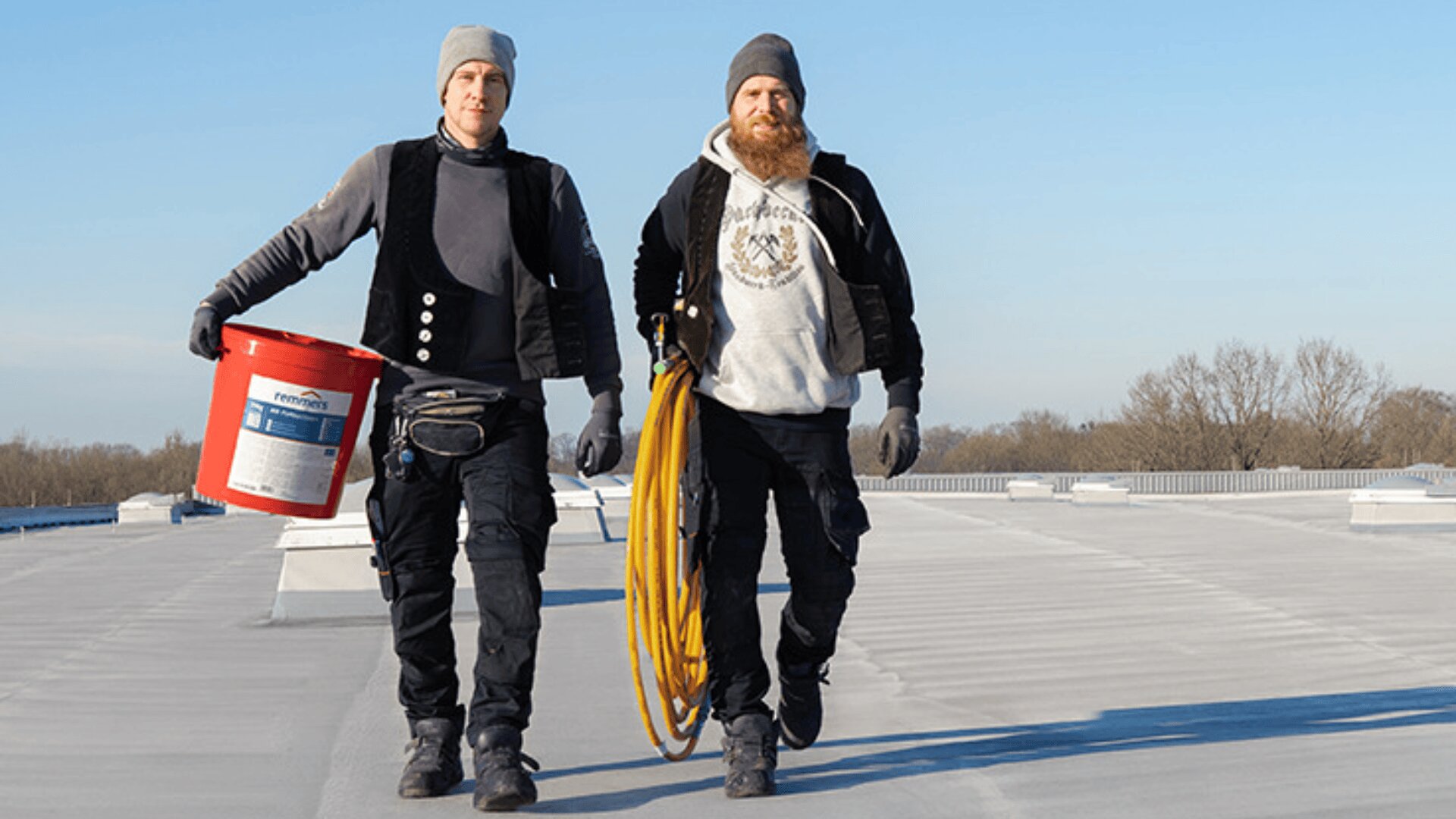 Zwei Arbeiter in Winterkleidung gehen auf einem Flachdach. Einer trägt einen roten Eimer, der andere einen gelben Schlauch. Beide tragen Handschuhe, Mützen und Jacken. Der Himmel ist klar und blau, im Hintergrund sind Bäume zu sehen.