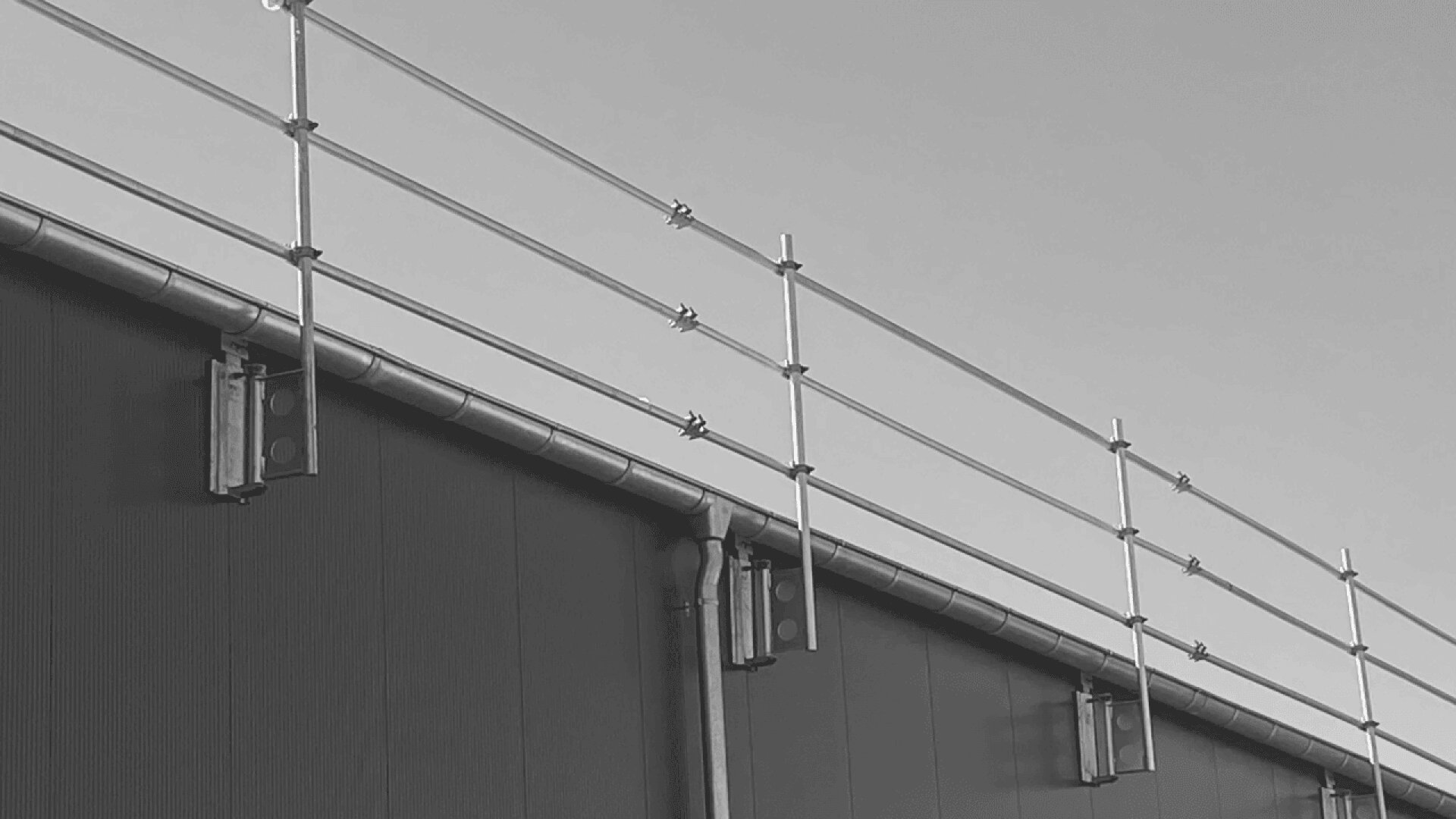 A black and white photo shows a rooftop safety railing system installed along the edge of a building, with vertical and horizontal metal bars silhouetted against the sky.