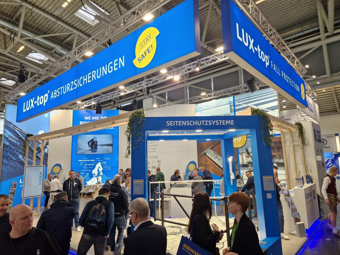 Ein belebter Messestand für LUX-top, der Absturzsicherungssysteme anbietet, mit Menschen, die miteinander interagieren, Bannern mit dem Markenzeichen des Unternehmens und Informationsdisplays unter einem großen blauen Überkopfschild.