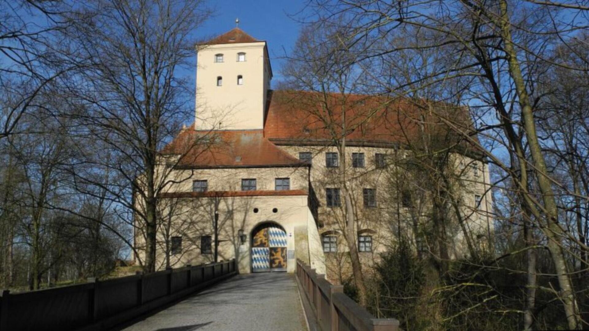 A historic castle with a tall square tower and red-tiled roof, surrounded by leafless trees. A bridge leads to an arched entrance decorated with blue and white diamond patterns. The sky is clear and blue.