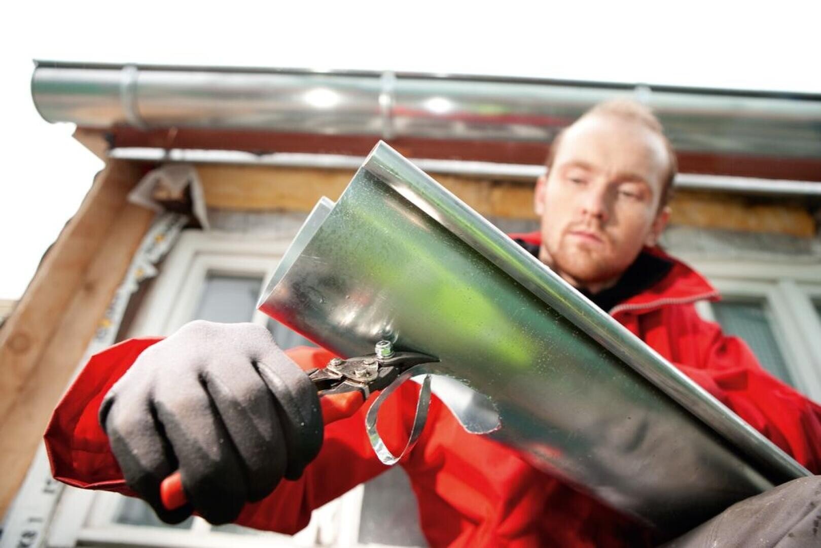 A man wearing gloves and a red jacket cuts off a piece of a shiny metal gutter with a pair of metal shears, while a house with a gutter system can be seen in the background.