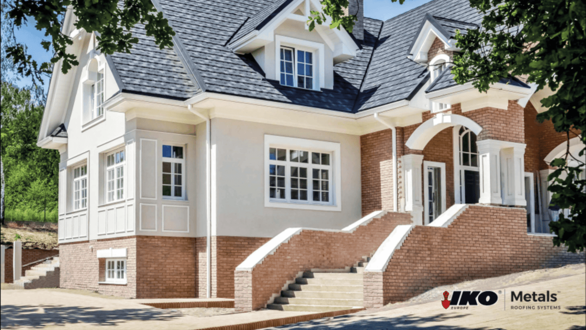 A modern two-story house with light brick and cream siding, large windows, a steep gray roof, and multiple gables. Steps lead to a white front door. The IKO Metals Roofing Systems logo is in the lower right corner.