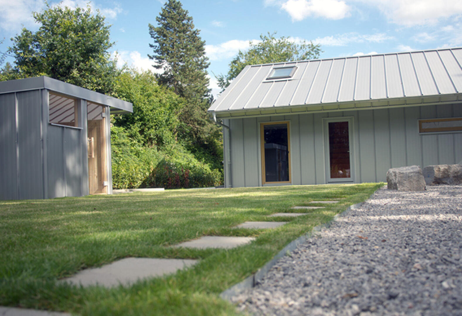 Moderner Hinterhof mit grünem Rasen, Steinpflasterweg, Kiesfläche und zwei grauen Metallgebäuden mit großen Fenstern, umgeben von Bäumen unter blauem Himmel.