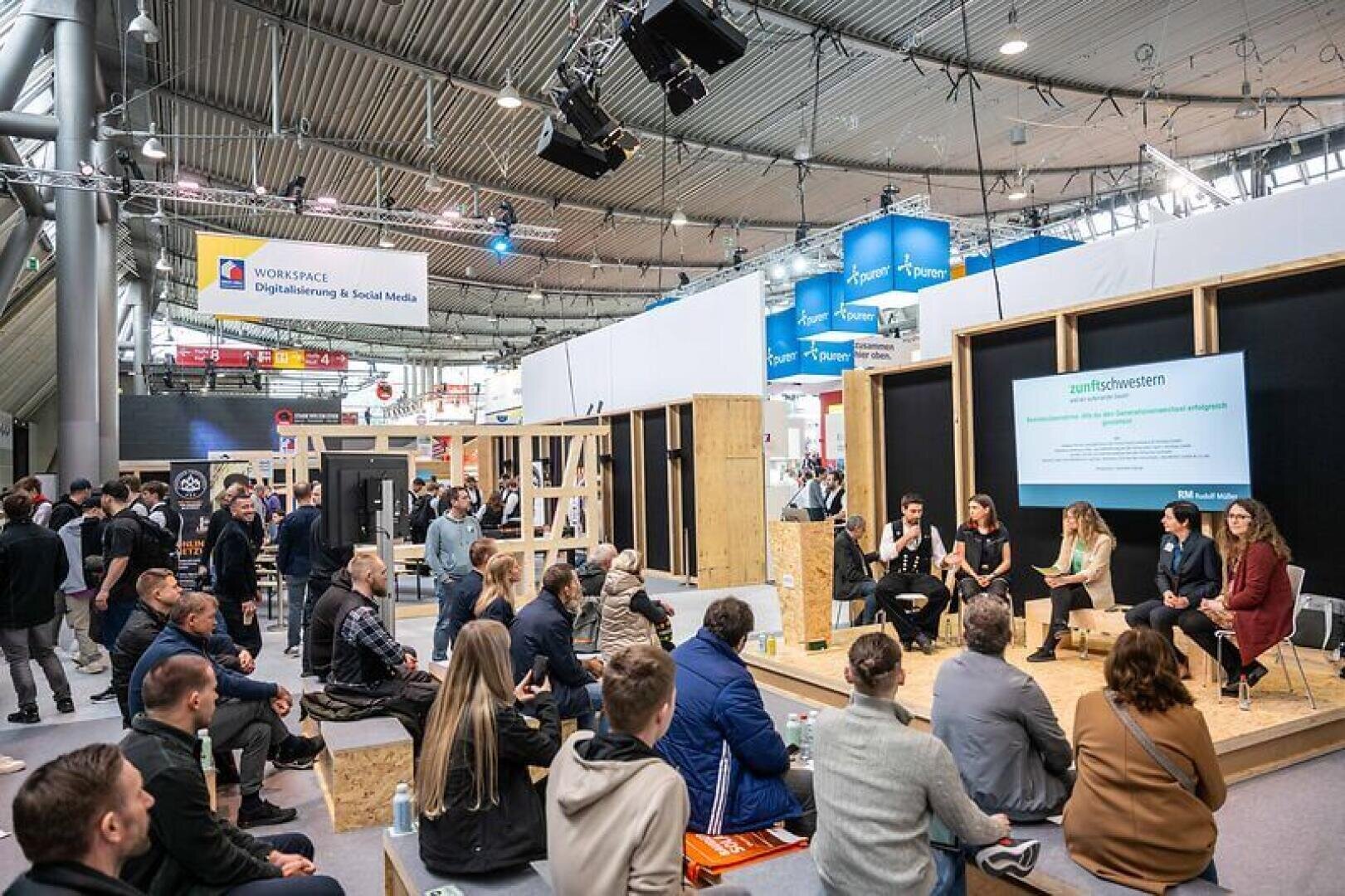 Eine Gruppe von Menschen sitzt auf Bänken und auf dem Boden und hört einer Podiumsdiskussion bei einer Indoor-Veranstaltung mit modernen Displays und digitaler Arbeitsplatzbeschilderung im Hintergrund zu.