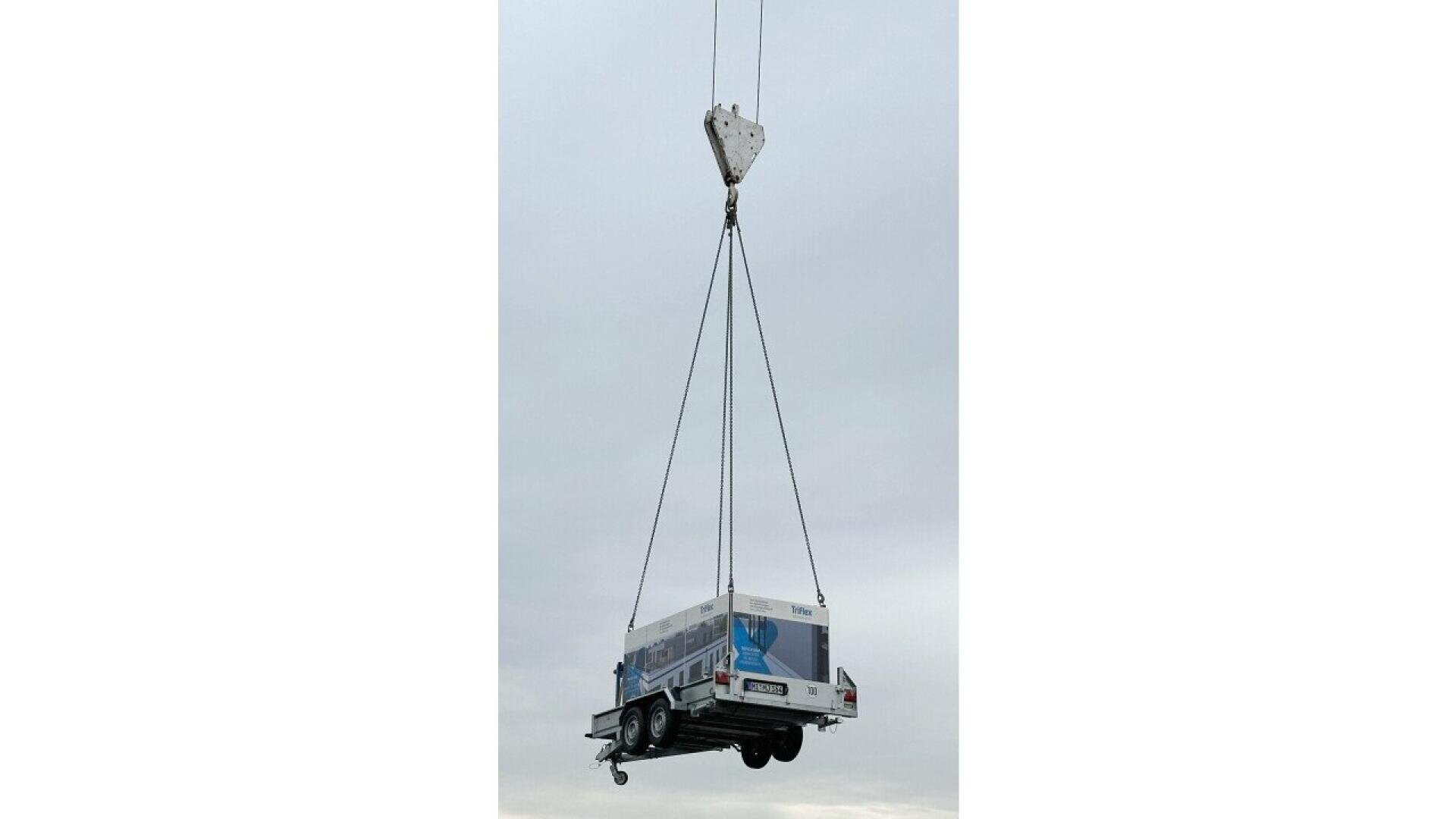 A utility truck is suspended in midair by four cables attached to a crane hook against a cloudy sky background.