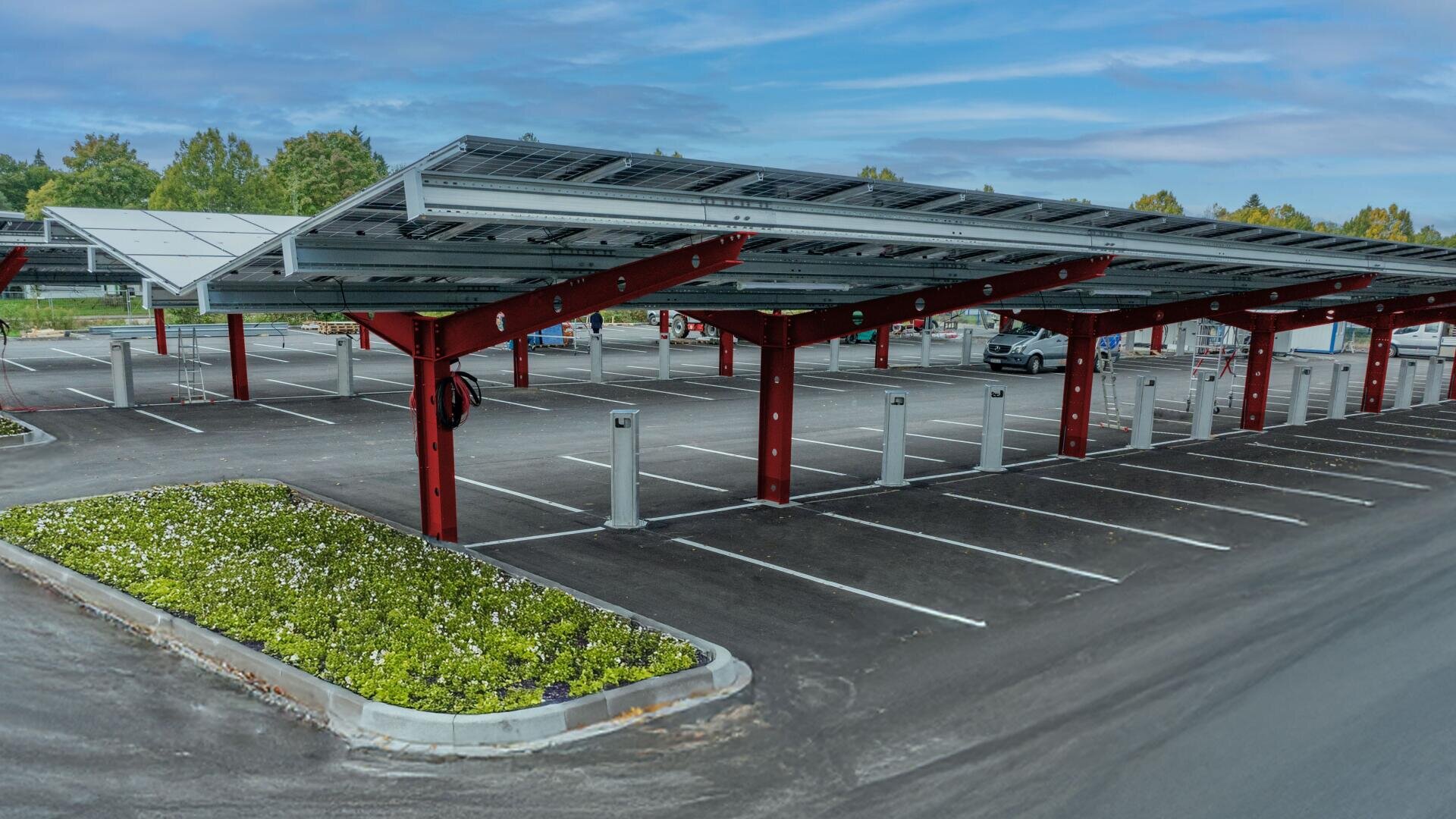 Ein Parkplatz mit überdachten Stellplätzen mit Solarzellen auf den Dächern und roten Metallstützen. Mehrere Ladestationen für Elektrofahrzeuge sind installiert, und es gibt einen begrünten Bereich mit Grünpflanzen.