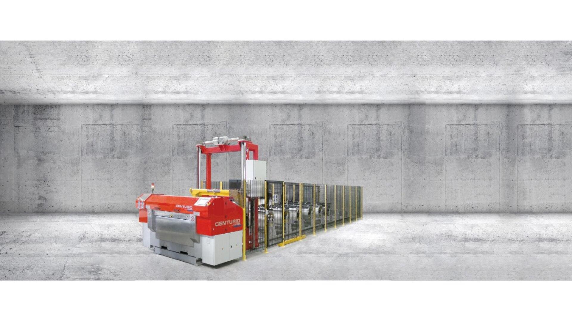 A large industrial machine with a red and white front and yellow railings stands in an empty, gray concrete room with a smooth floor and textured walls.