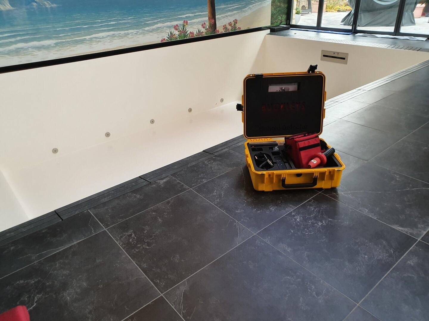 A yellow hard case containing various tools and equipment stands open on a dark tiled floor next to an empty indoor swimming pool with a mural of a beach scene on the wall.