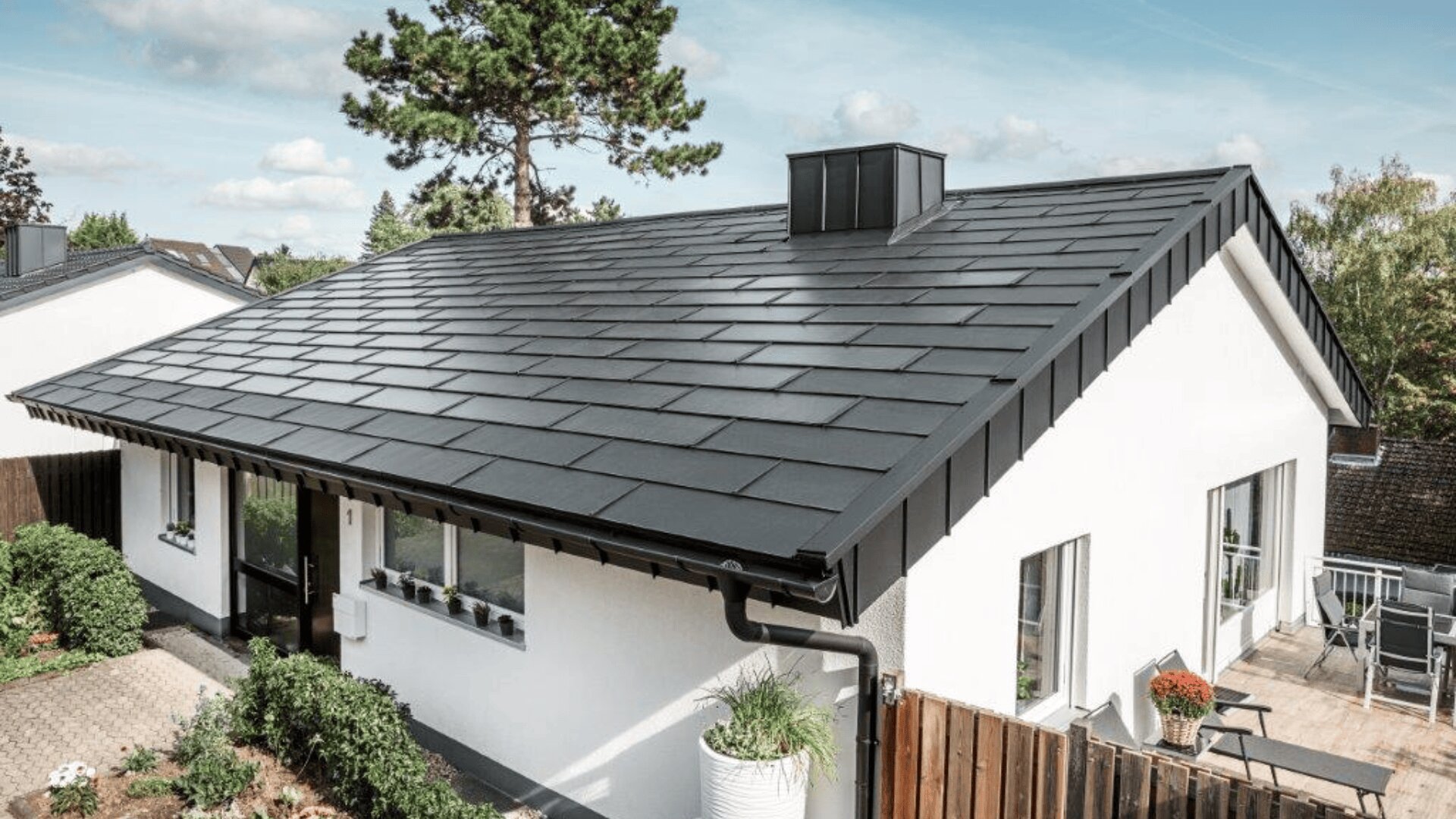 Ein modernes weißes Haus mit großen Fenstern und einem dunklen, schrägen Metalldach mit zentraler Lüftungsöffnung. Das Haus verfügt über einen kleinen Garten, Topfpflanzen in der Nähe des Eingangs und ist von Bäumen umgeben, die von einem teilweise bewölkten Himmel umgeben sind.