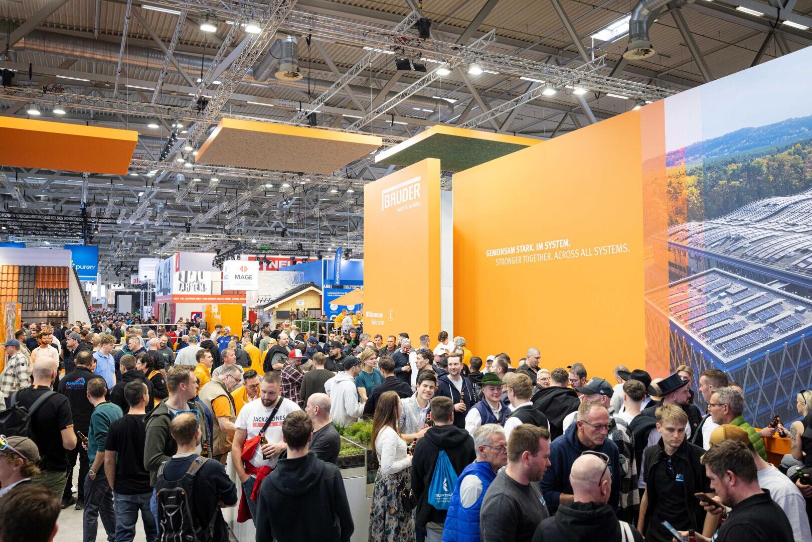 Eine große Menschenmenge besucht eine Indoor-Messe oder -Ausstellung, versammelt um leuchtend orangefarbene Stände bei heller Beleuchtung, mit verschiedenen Displays und Schildern, die in der geräumigen Halle sichtbar sind.