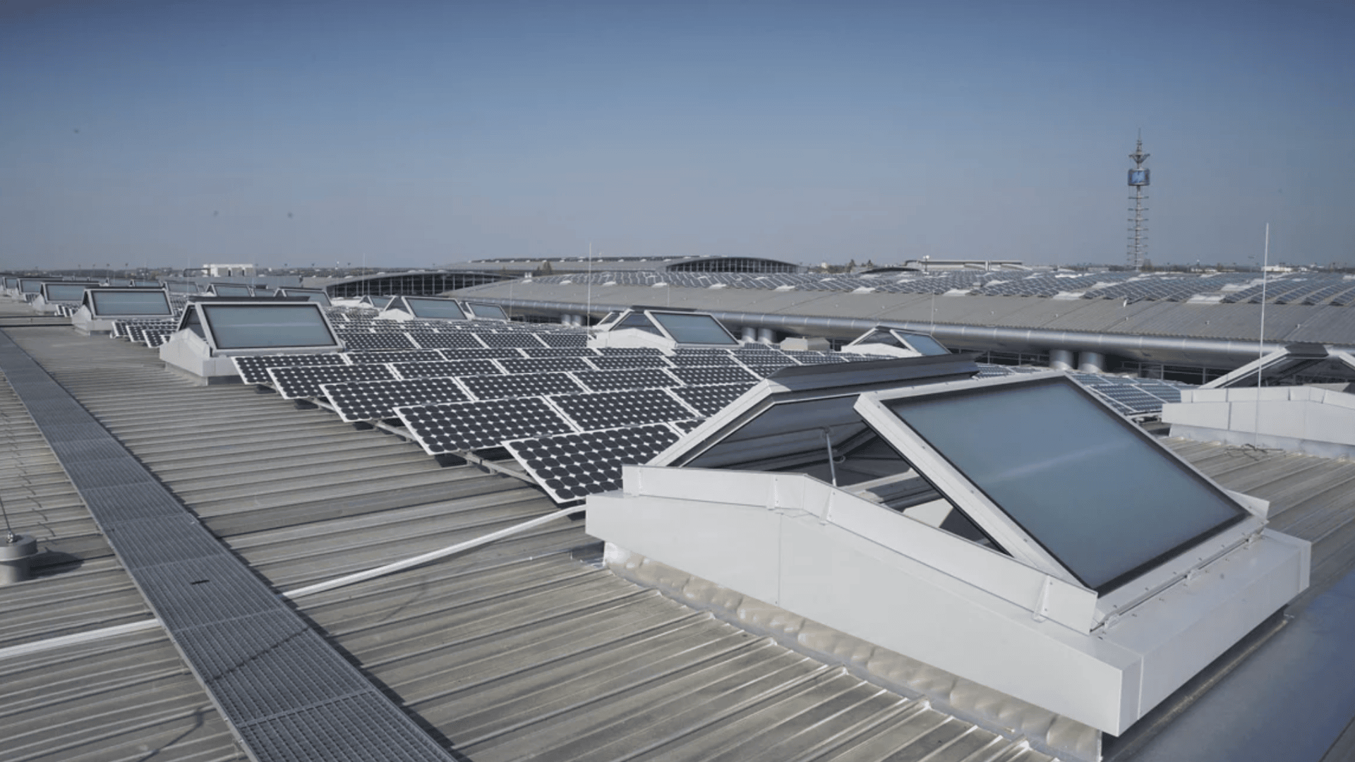 Auf einem großen industriellen Metalldach sind unter einem klaren blauen Himmel Reihen von Solarmodulen installiert; Fenster sind in die Dachkonstruktion integriert.