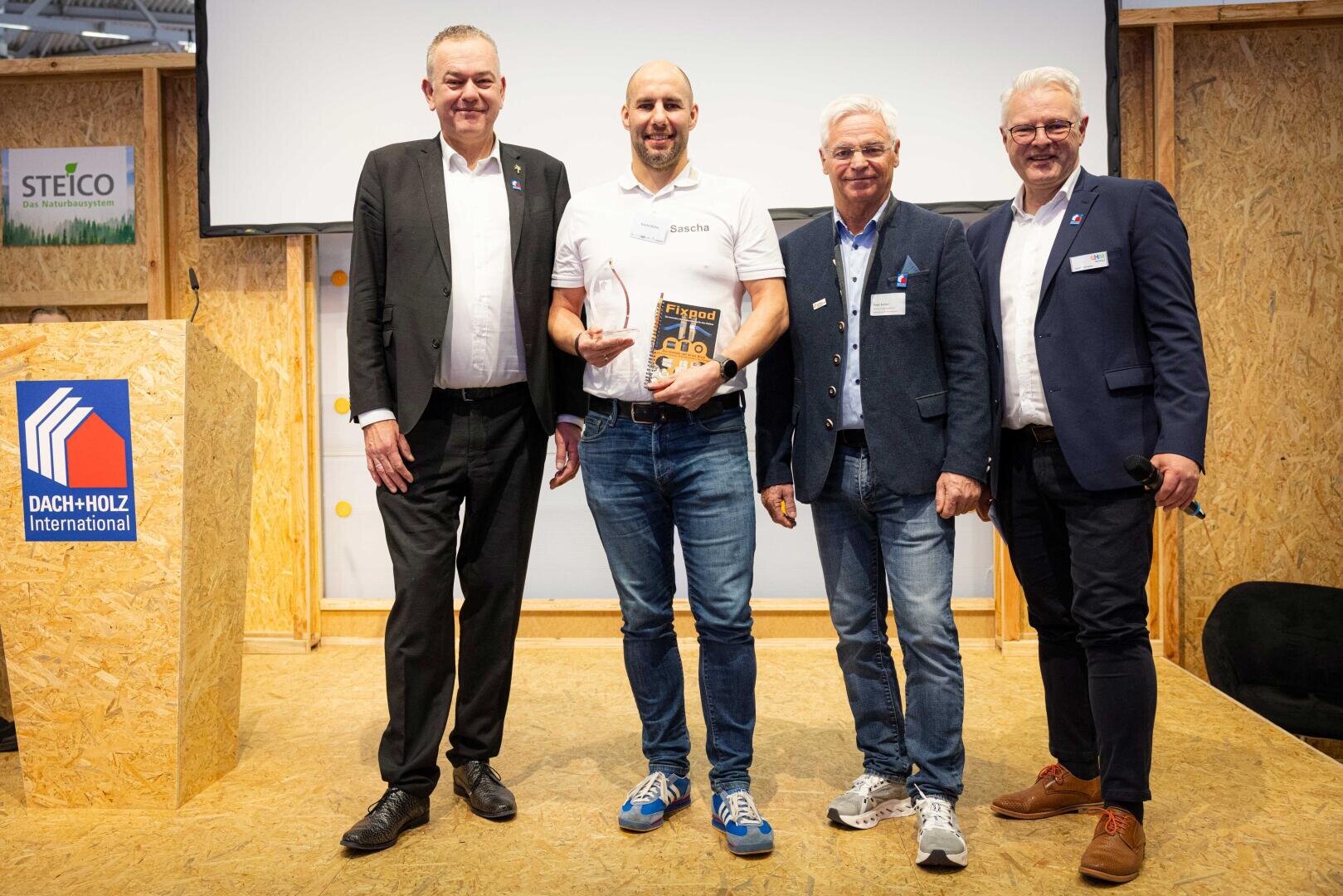 Vier Männer stehen auf einer Bühne, einer hält einen gläsernen Preis und ein Buch in der Hand. Sie sind in Business- und Freizeitkleidung gekleidet und lächeln in die Kamera. Ein Schild von Dach+Holz International ist auf dem Podium in der Nähe zu sehen.