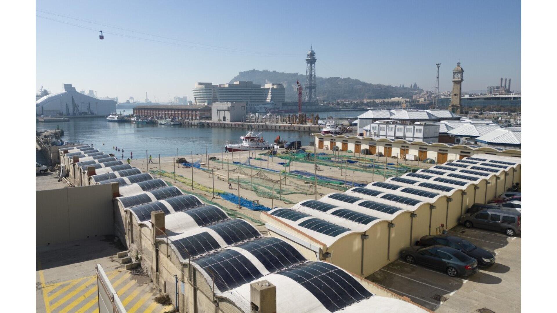 Rows of industrial buildings with curved roofs covered in solar panels near a waterfront; boats and city structures are visible in the background under a clear blue sky.