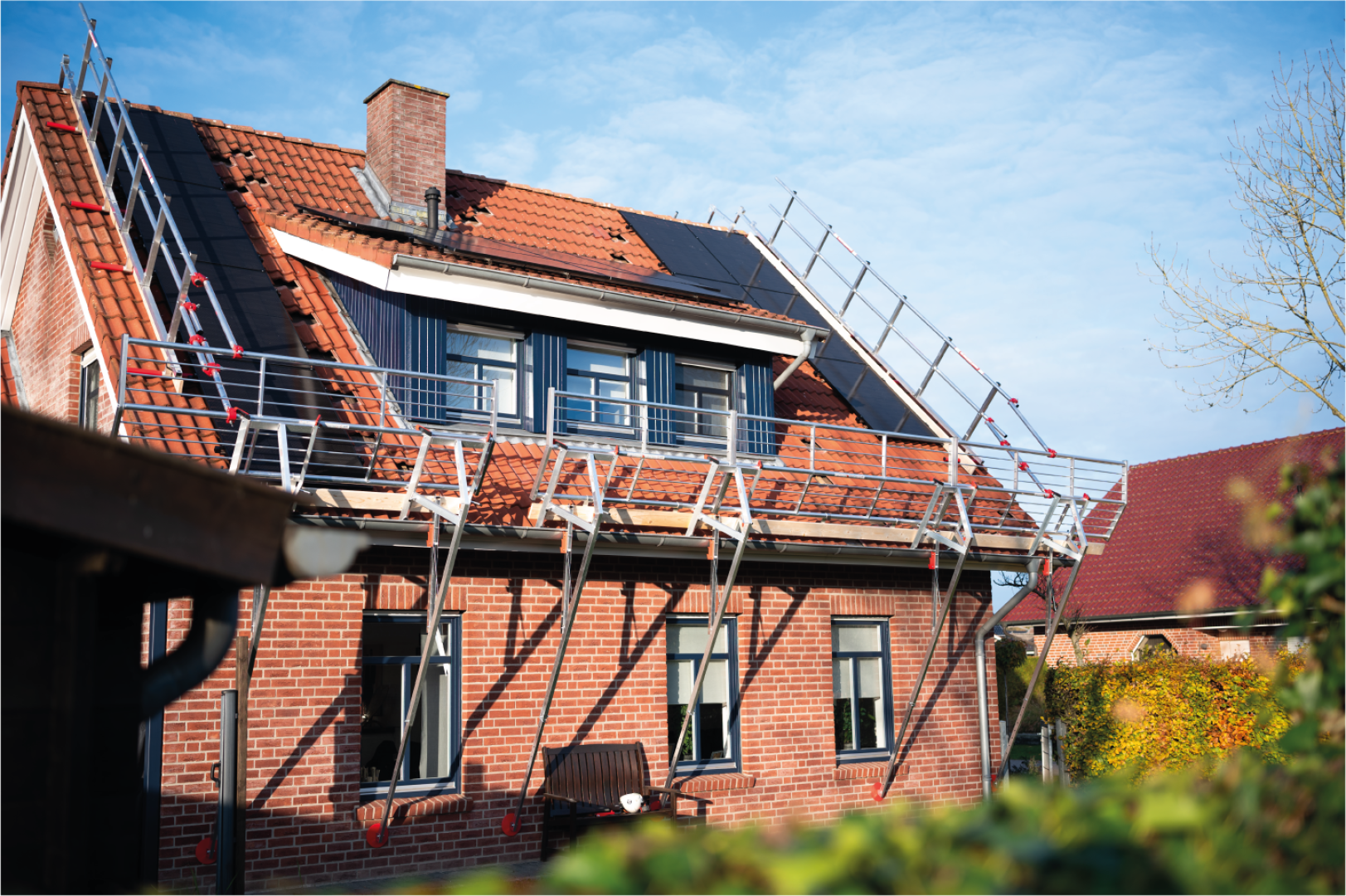 Ein Backsteinhaus mit einem Gerüst auf dem Ziegeldach, auf dem einige Ziegel fehlen. Es sind Sonnenkollektoren installiert und ein Schornstein ist sichtbar; der Himmel ist klar und blau.