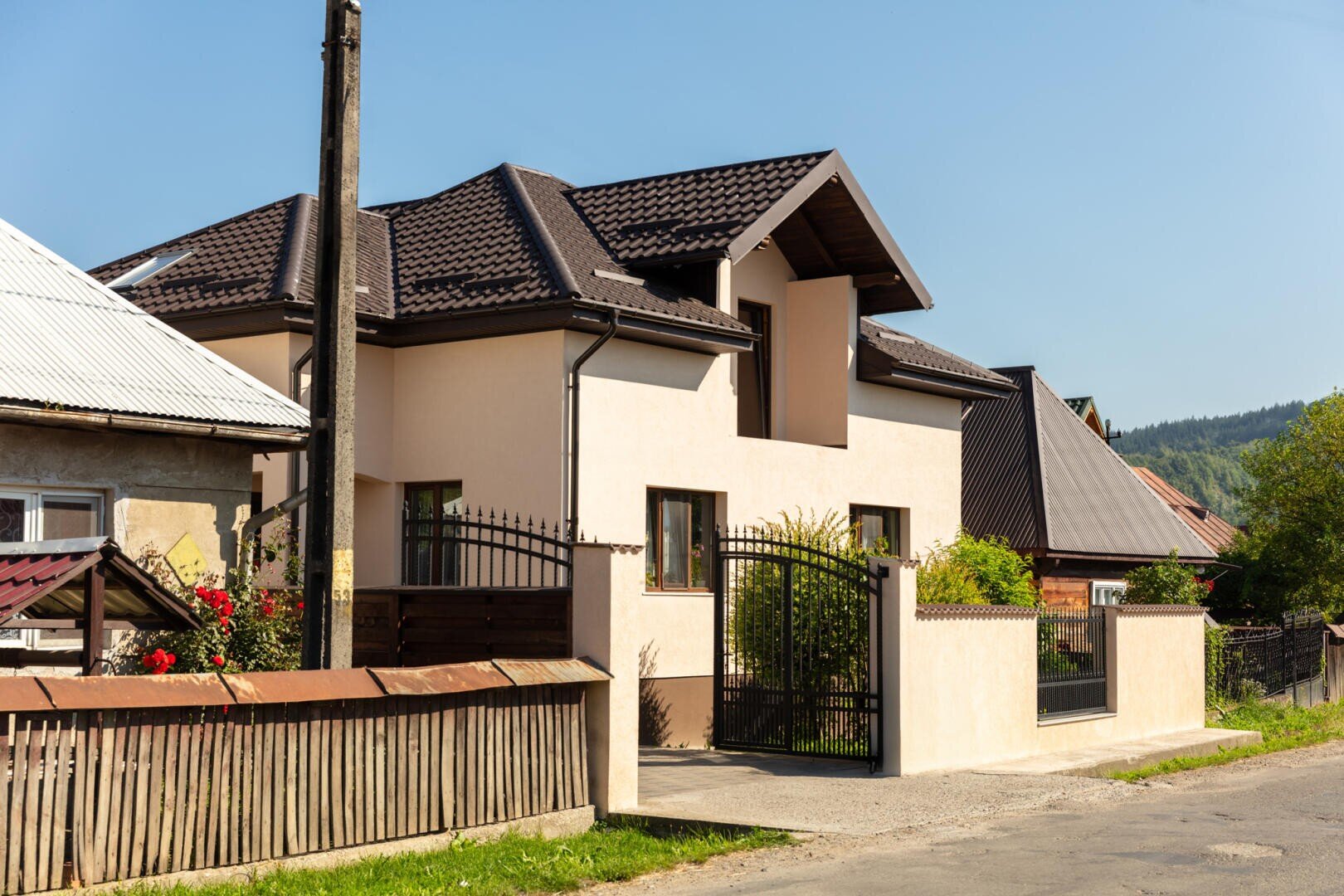 A modern beige house with a dark brown roof and a black metal gate, surrounded by a low cream-colored wall, next to an older wooden fence and other houses on a sunny day. Trees and hills can be seen in the background.