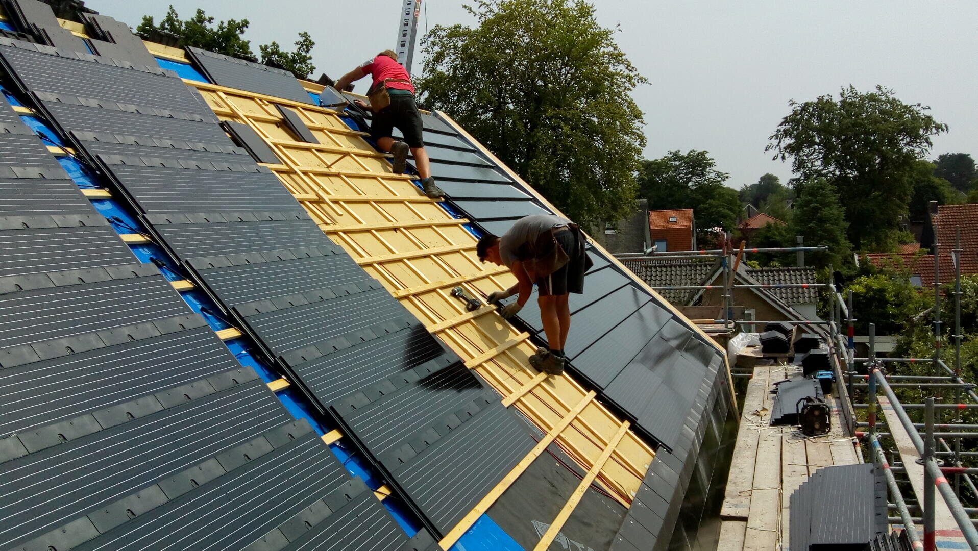 Zwei Arbeiter auf einem Gerüst installieren Solarmodule auf einem Schrägdach und befestigen die schwarzen Module auf einem Holzgitter. An einem klaren Tag sind im Hintergrund Bäume und Dächer zu sehen.