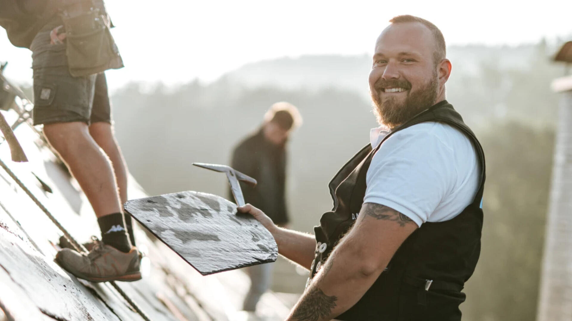 Ein lächelnder Bauarbeiter mit Bart hält einen Dachziegel und ein Werkzeug in der Hand, während er an einem Schrägdach arbeitet. Die Beine eines weiteren Arbeiters sind sichtbar, eine dritte Person ist im Hintergrund verschwommen. Die Szene spielt im Freien bei Tageslicht.