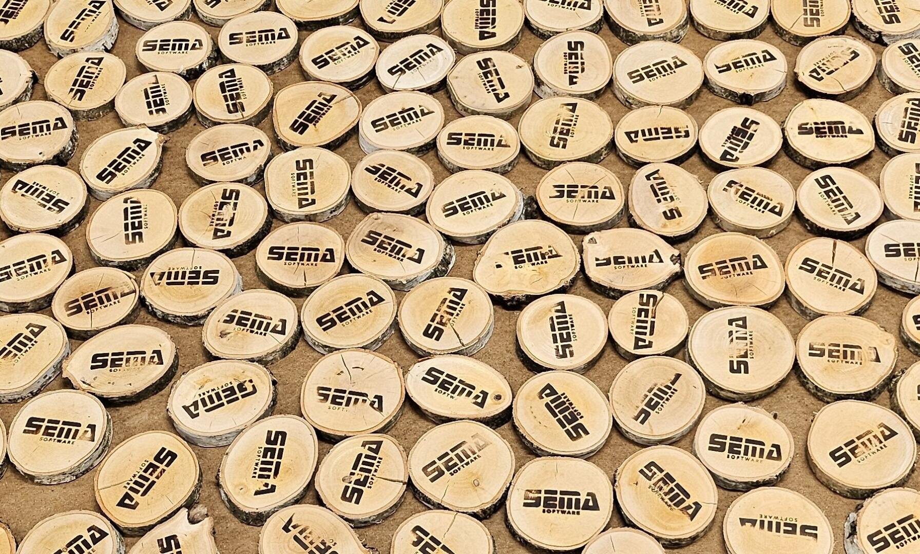 Dutzende runde Holzscheiben mit den eingebrannten Worten SEMA und SOFTBALL liegen leicht überlappend auf einer sandigen Fläche.