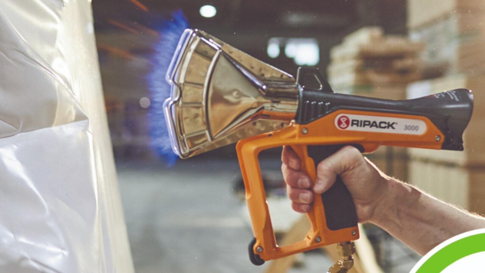 A person uses an orange and black RIPACK 3000 heat gun, emitting a blue flame, to shrink a large sheet of plastic in an industrial warehouse setting.
