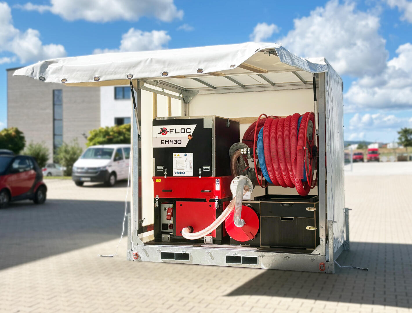 An X-Floc EM430 blow-in machine is mounted in an open trailer with a hose reel and red hoses. The platform is located on a paved area with buildings and vehicles in the background.