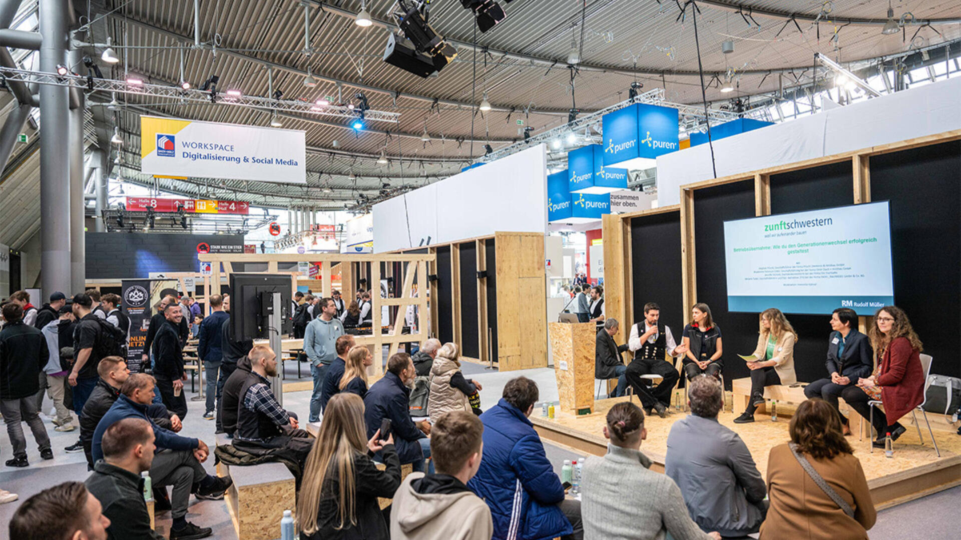 Eine Gruppe von Menschen sitzt und hört einer Podiumsdiskussion bei einer Technologie- oder Geschäftsveranstaltung in einem großen, modernen Veranstaltungsort mit digitalen Bildschirmen und verschiedenen Ständen im Hintergrund zu.