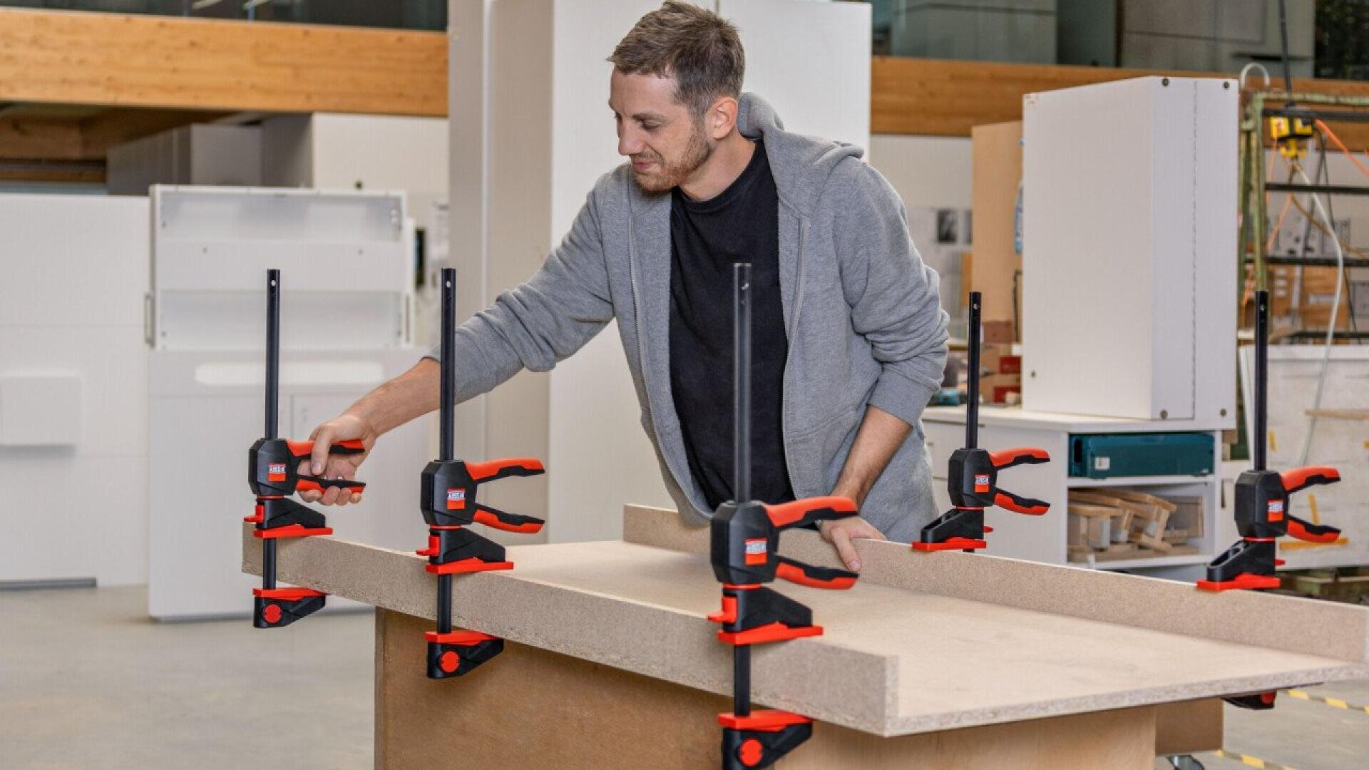 A man in a gray hoodie uses several red and black clamps to secure two pieces of wood together in a workshop setting.