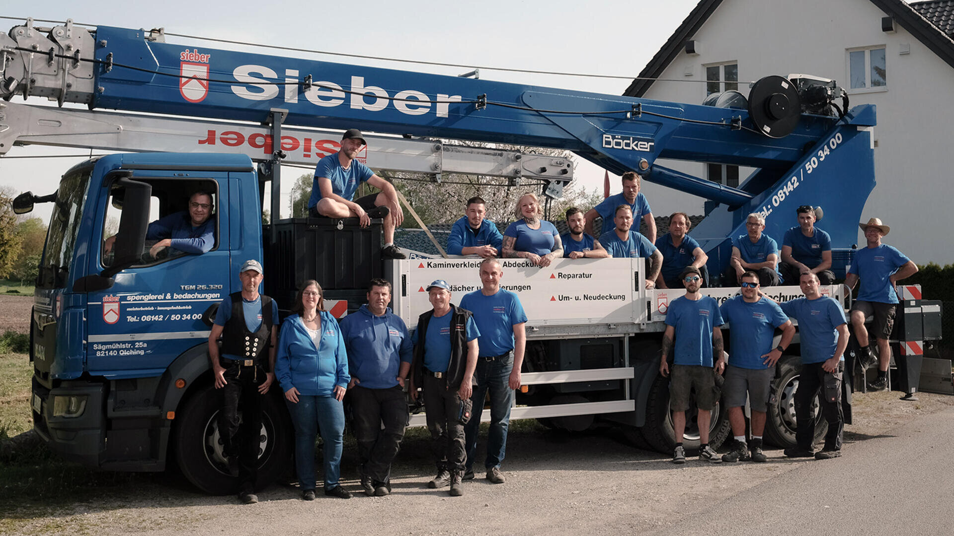Eine Gruppe von 18 Personen in blauen Hemden posiert an einem sonnigen Tag vor einem großen blauen Sieber-LKW mit Kran, der in der Nähe eines weißen Hauses geparkt ist. Einige stehen, andere sitzen auf dem LKW, alle lächeln in die Kamera.