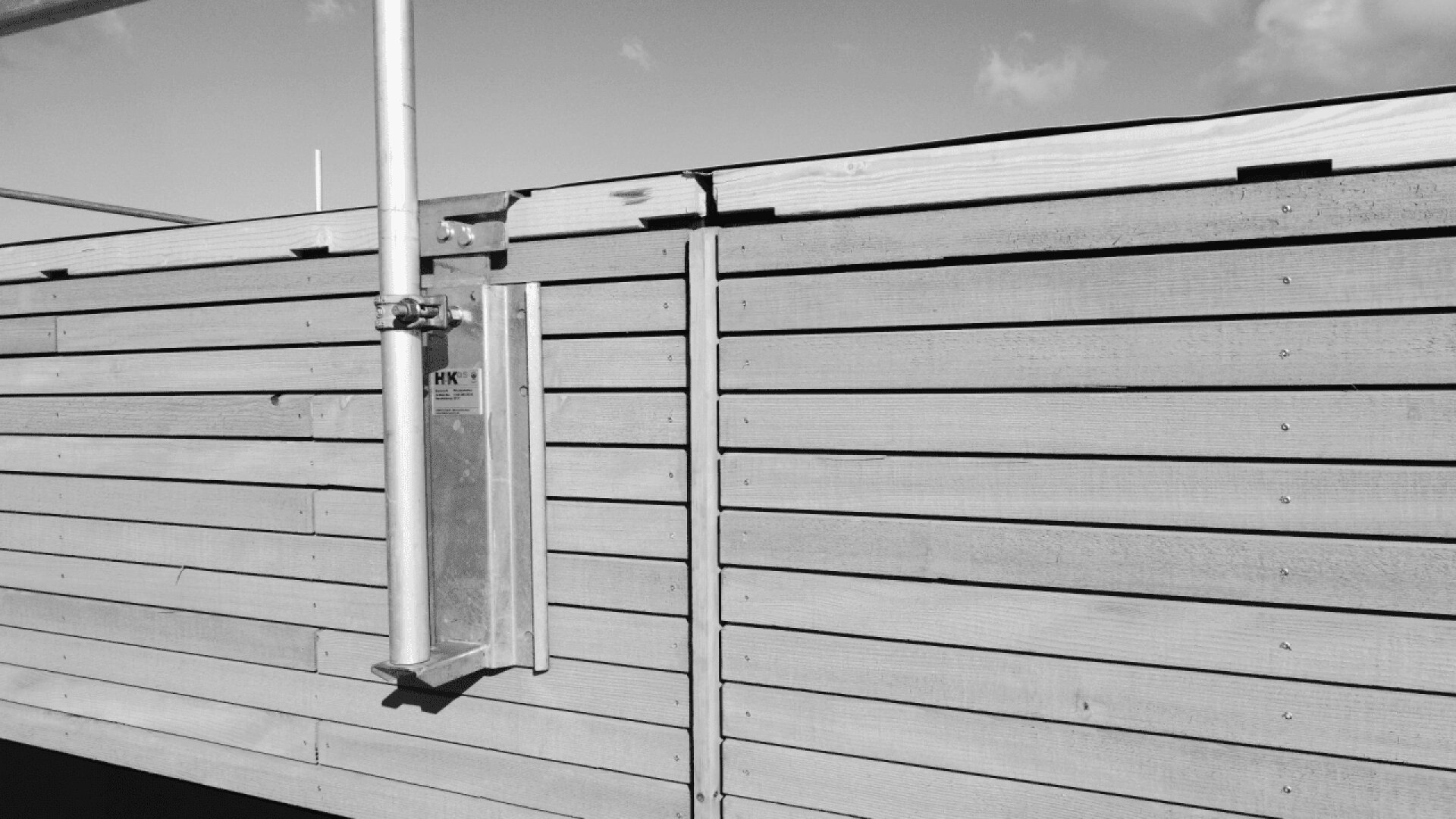 A metal utility box is mounted on a wooden fence made of horizontal planks, with a metal pipe extending upward on the left side. The image is in black and white under a partly cloudy sky.