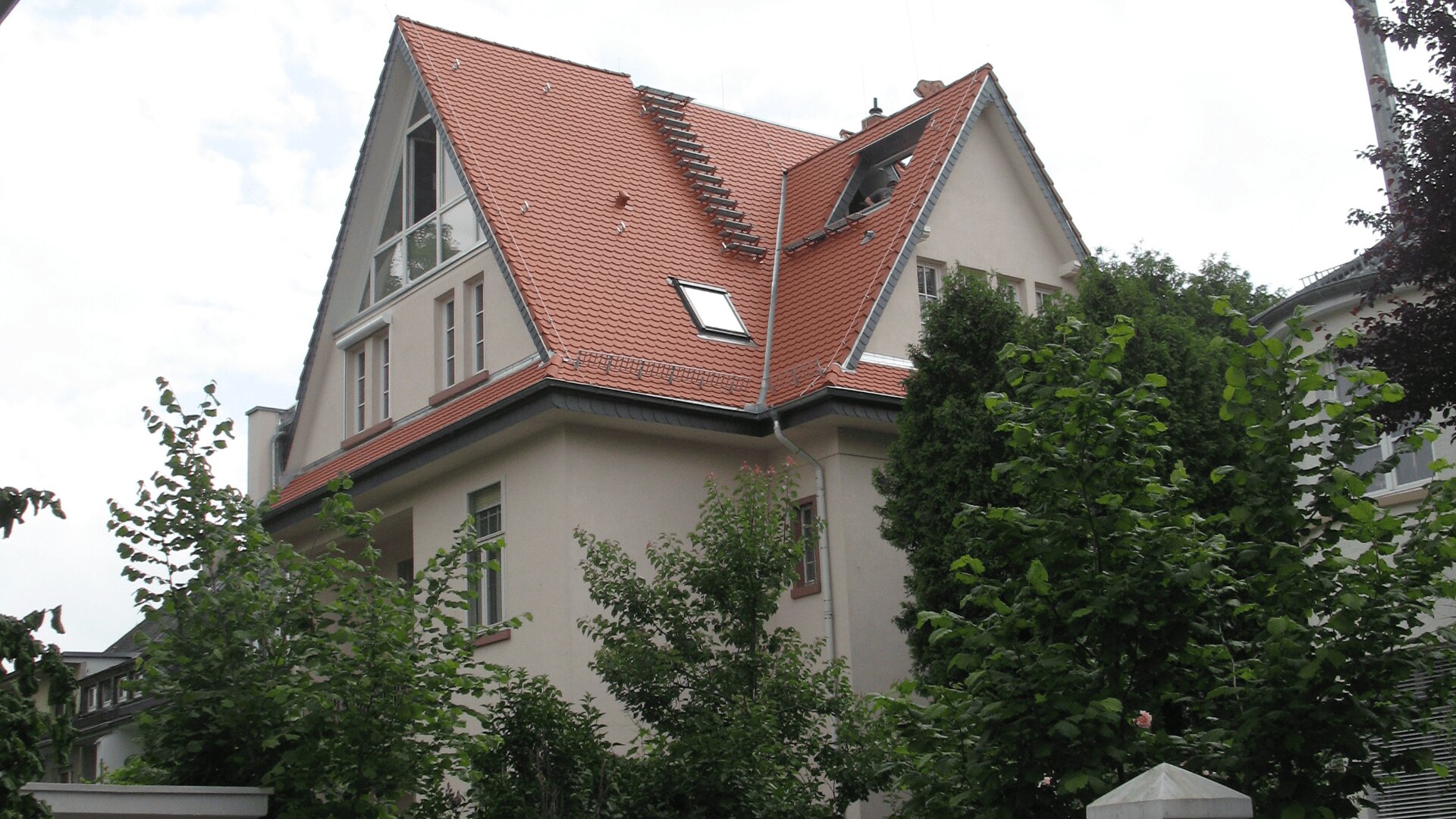 Ein zweistöckiges Haus mit hellen Wänden und einem steilen roten Ziegeldach, mehreren Fenstern und umgeben von grünen Bäumen.
