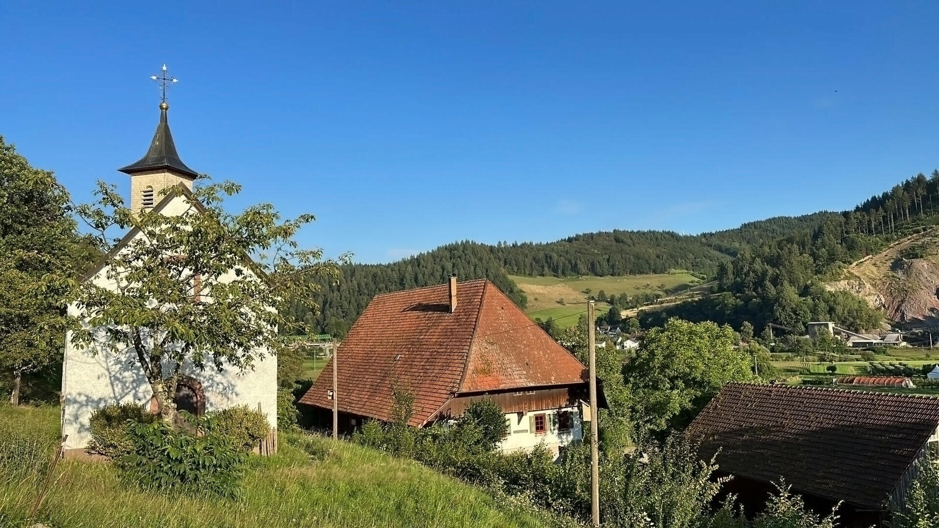 Eine kleine weiße Kirche mit einem Kirchturm steht neben Häusern mit roten Ziegeldächern, umgeben von Grün und Bäumen, vor einer Kulisse bewaldeter Hügel unter einem klaren blauen Himmel.