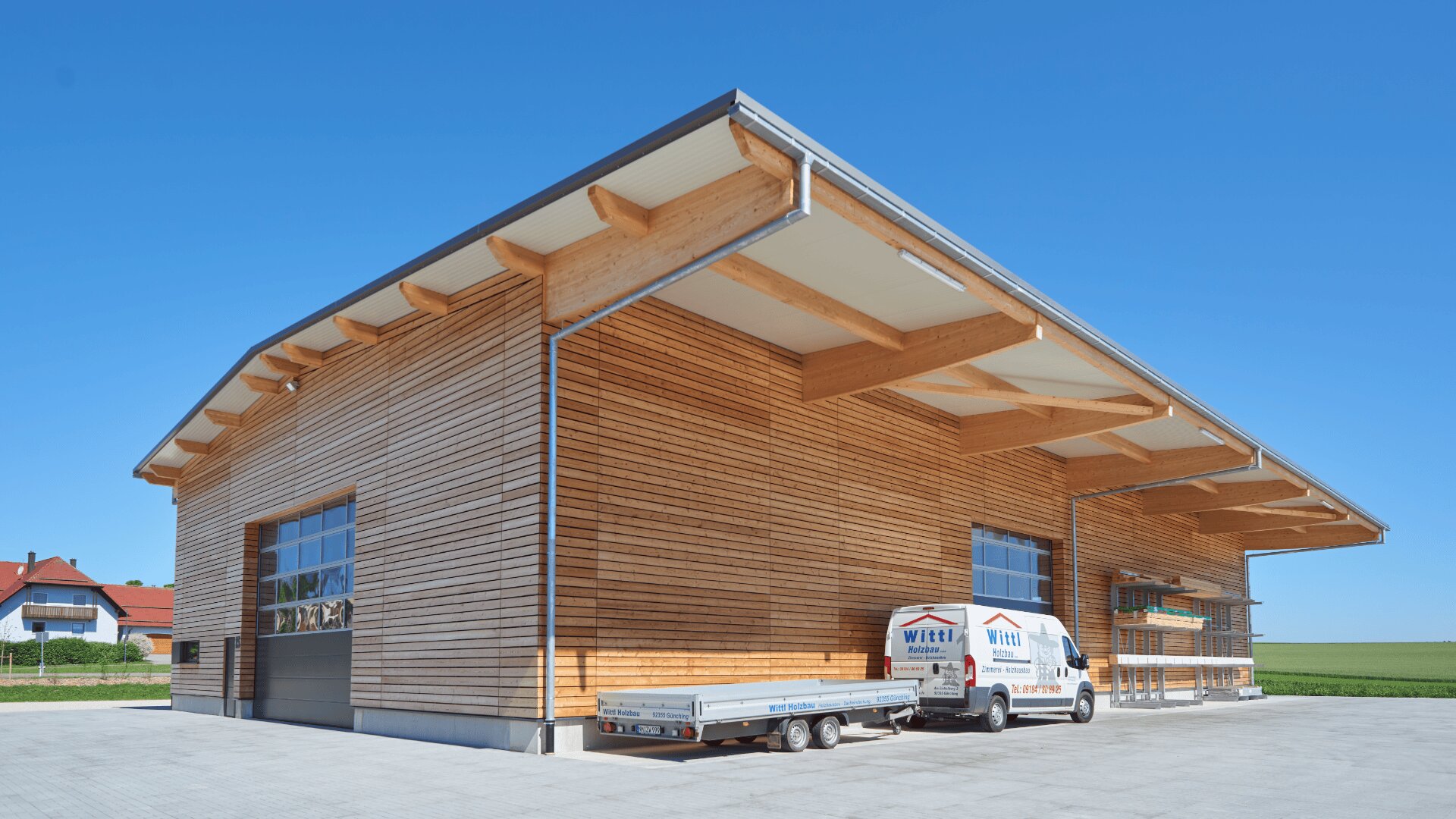 Ein modernes Industriegebäude aus Holz mit großen Fenstern und einem Metalldach. Ein weißer Lieferwagen und ein Anhänger stehen draußen auf einem gepflasterten Platz, im Hintergrund der strahlend blaue Himmel.