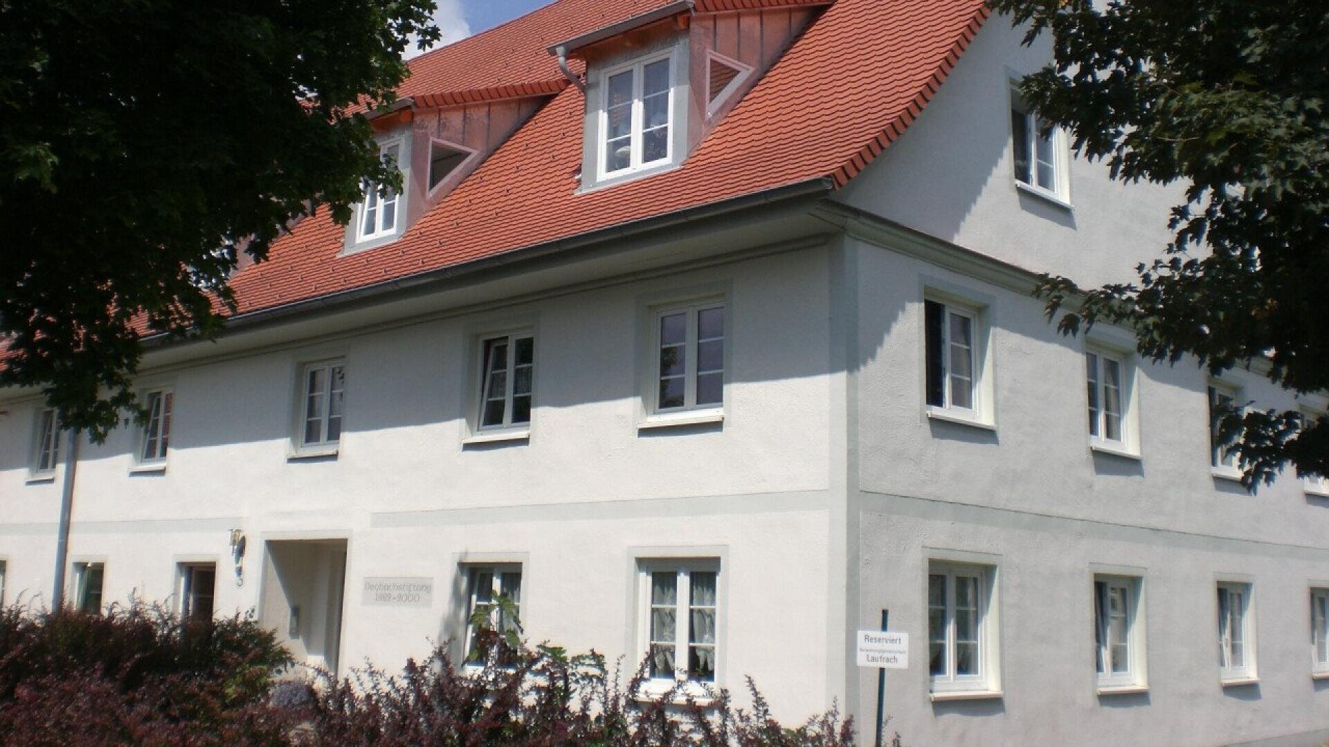 A white two-story building with a red-tiled roof and multiple windows, surrounded by trees and bushes, under a clear sky.