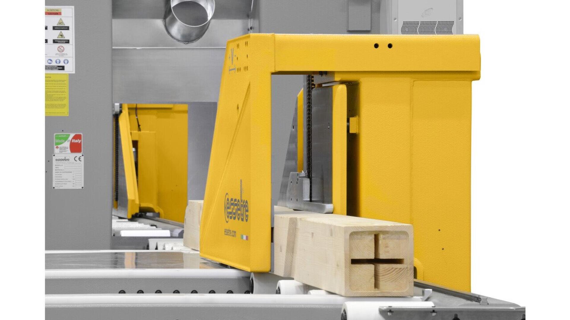 A yellow industrial woodworking machine is cutting a rectangular wooden beam with rollers guiding the wood. The beam has a cross-shaped cut on its end. Various safety and instruction labels are visible on the machine.