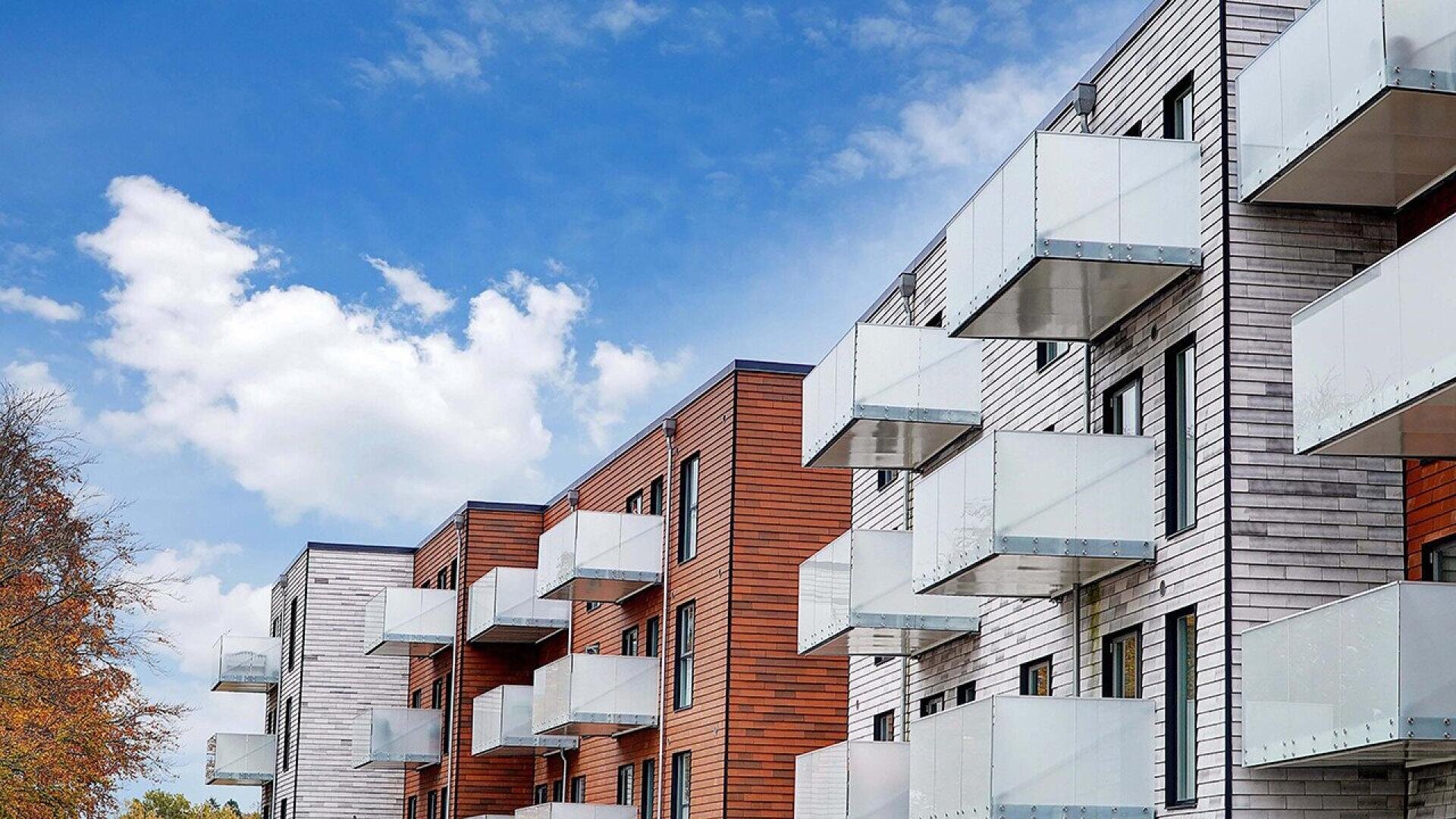 Modern apartment building with wooden and white panel exteriors, featuring multiple glass balconies, under a blue sky with white clouds. Trees with autumn-colored leaves are visible on the left.