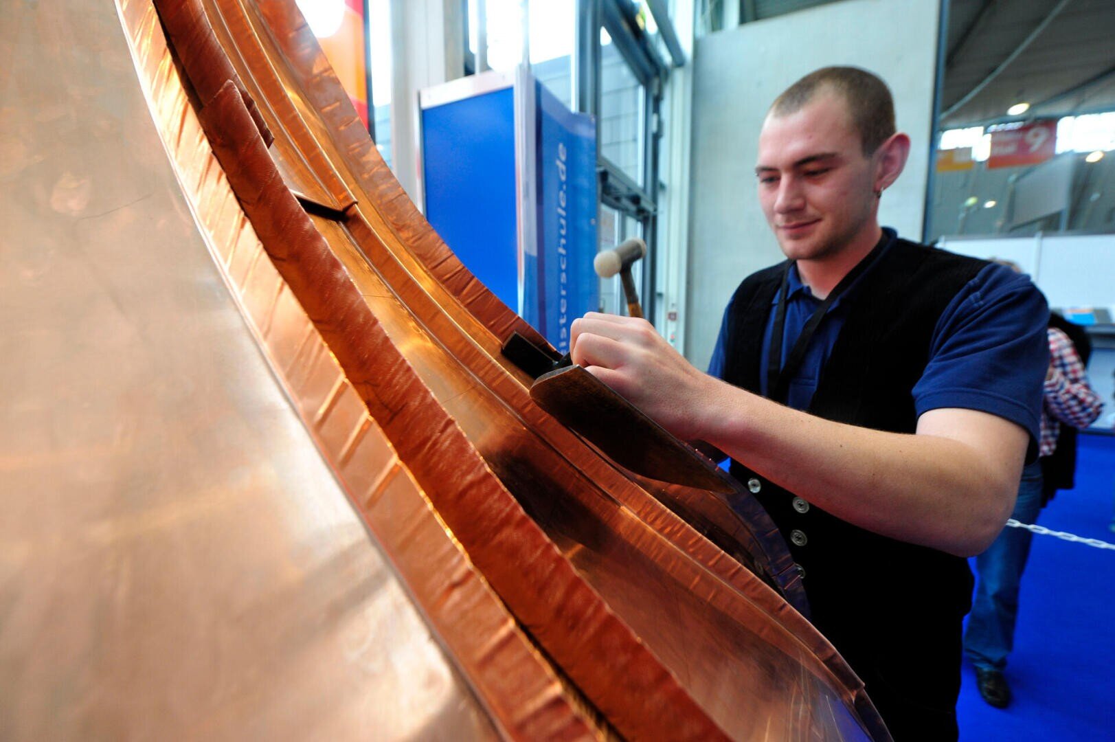 A man wearing a dark vest uses a small hammer to shape or work on a large, curved sheet of metal, possibly copper, at an indoor event or exhibition. Blue carpeting and display boards are in the background.