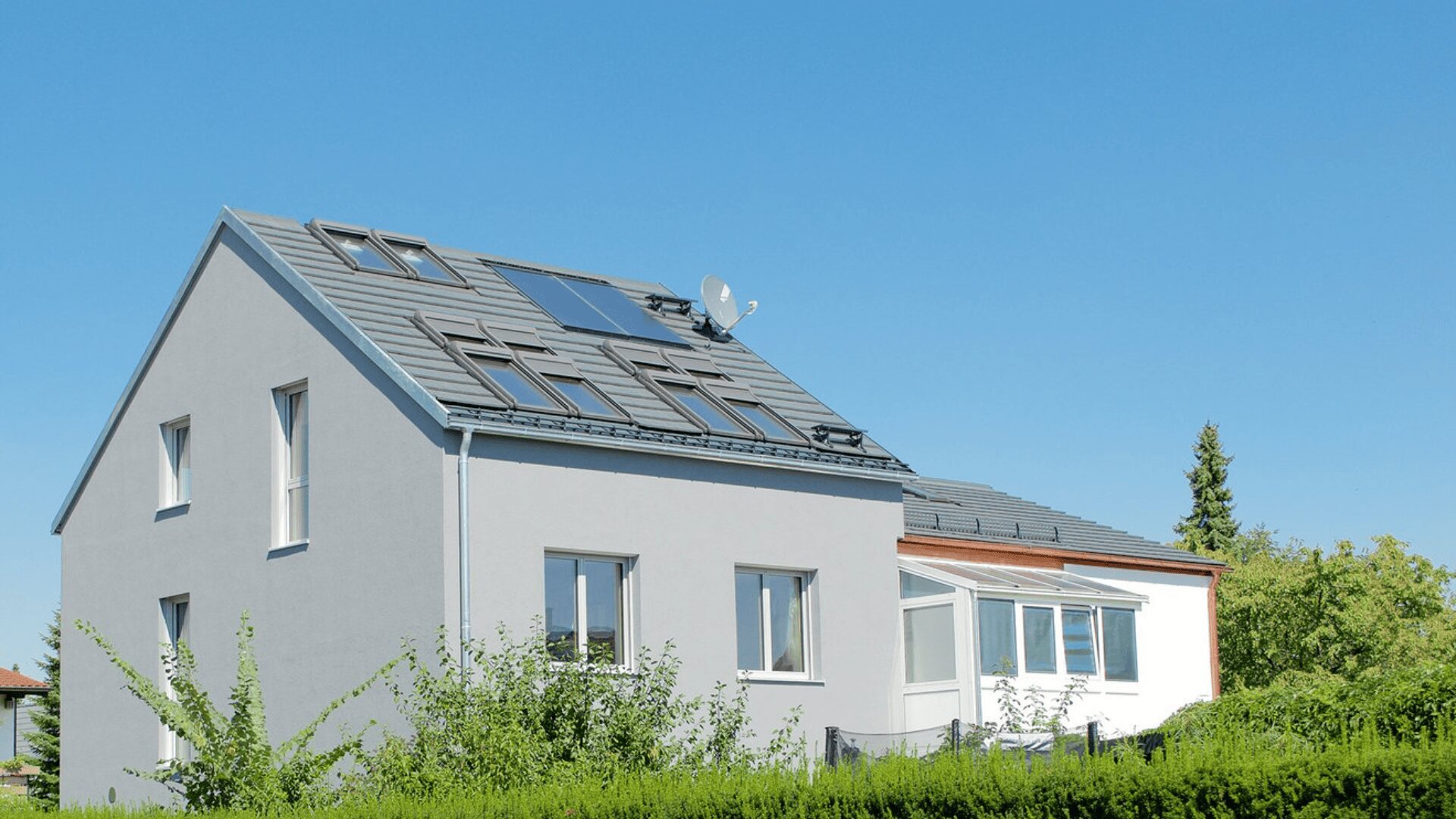 Ein modernes zweistöckiges Haus mit hellgrauer Fassade, großen Fenstern, Solarmodulen und einer Satellitenschüssel auf dem Dach. Ein Wintergarten ist angebaut, und das Haus ist von viel Grün umgeben, unter einem strahlend blauen Himmel.