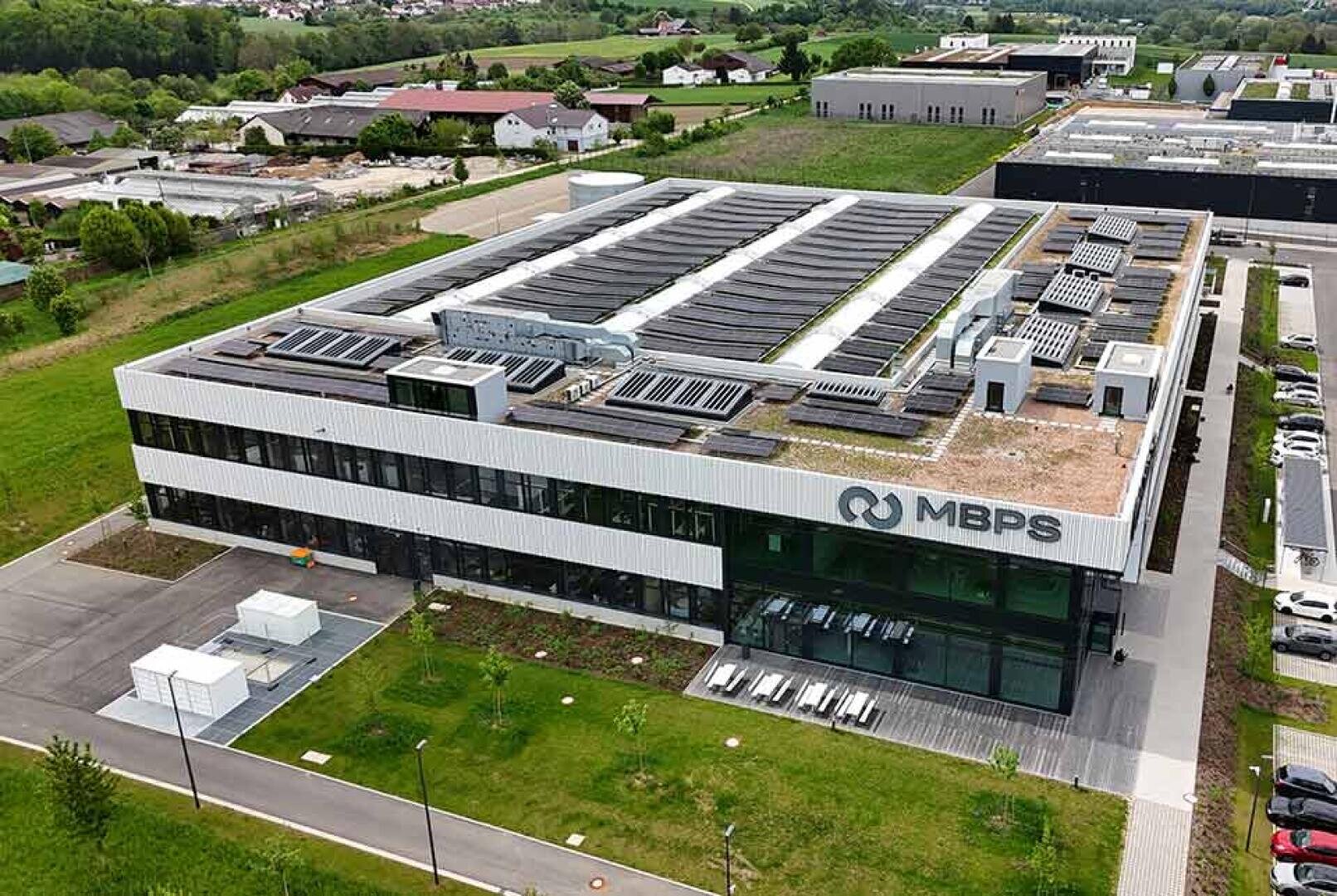 Aerial view of a modern two-story building with MBPS logo on the facade, solar panels and green roof, surrounded by grass, trees and other buildings in a semi-rural area.