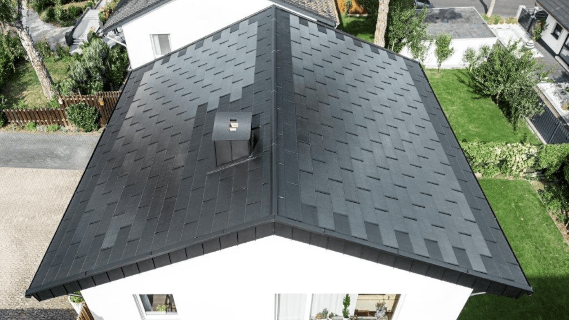 Luftaufnahme eines modernen Hauses mit einem dunkelgrauen, ordentlich gedeckten Satteldach mit einem kleinen quadratischen Schornstein in der Mitte, umgeben von grünen Rasenflächen und Bäumen.