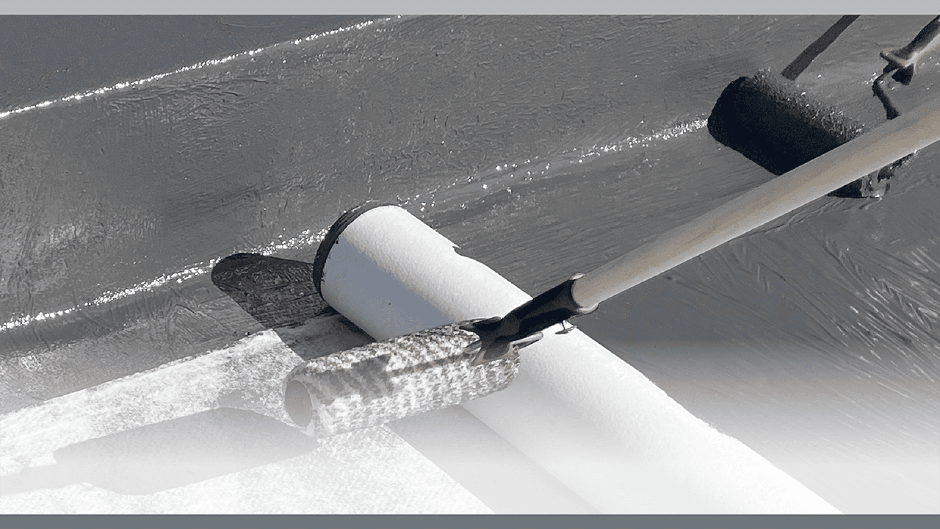 A paint roller with a long handle is being used to apply a white coating to a dark, flat surface, possibly a roof or floor, with another roller seen in the background.