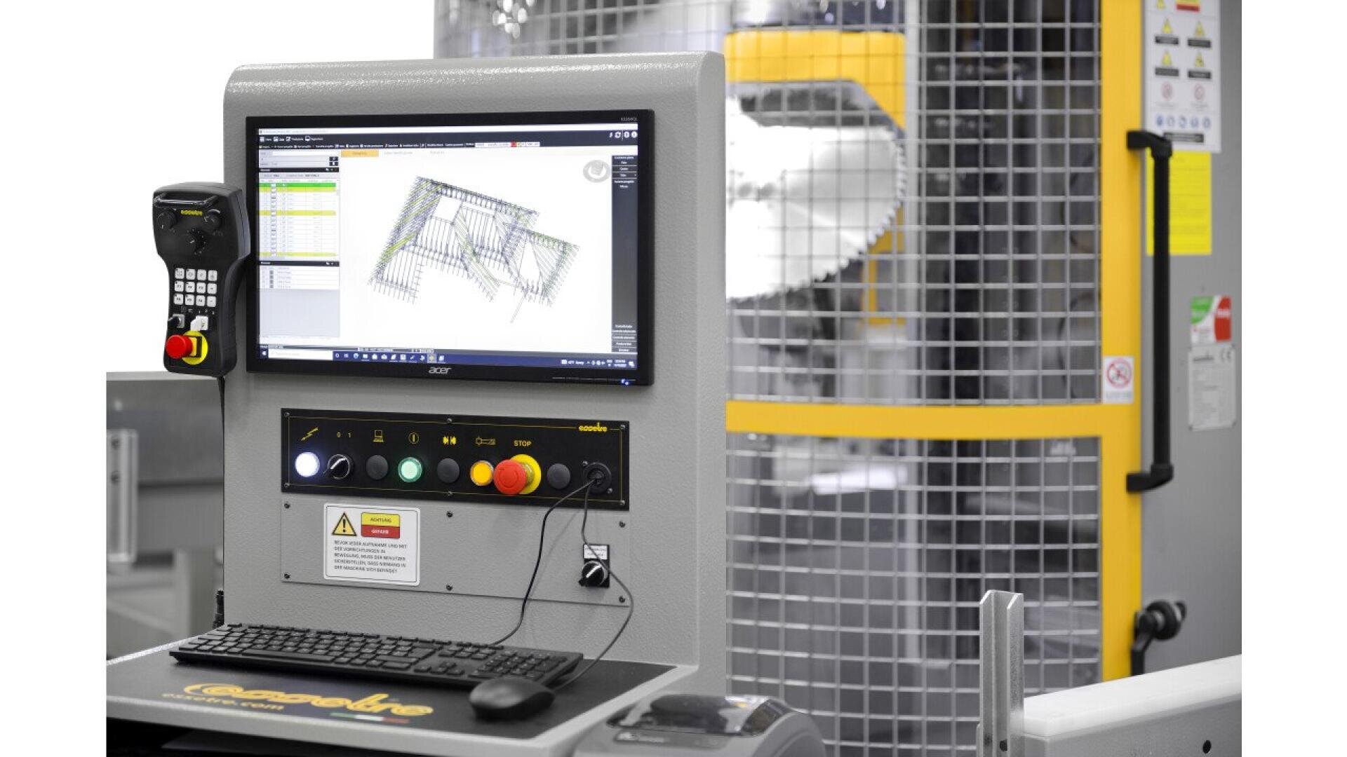 A close-up of an industrial machine control panel with a monitor displaying a technical diagram, several buttons, a keyboard, and safety signage. A yellow and gray fenced machine is visible in the background.