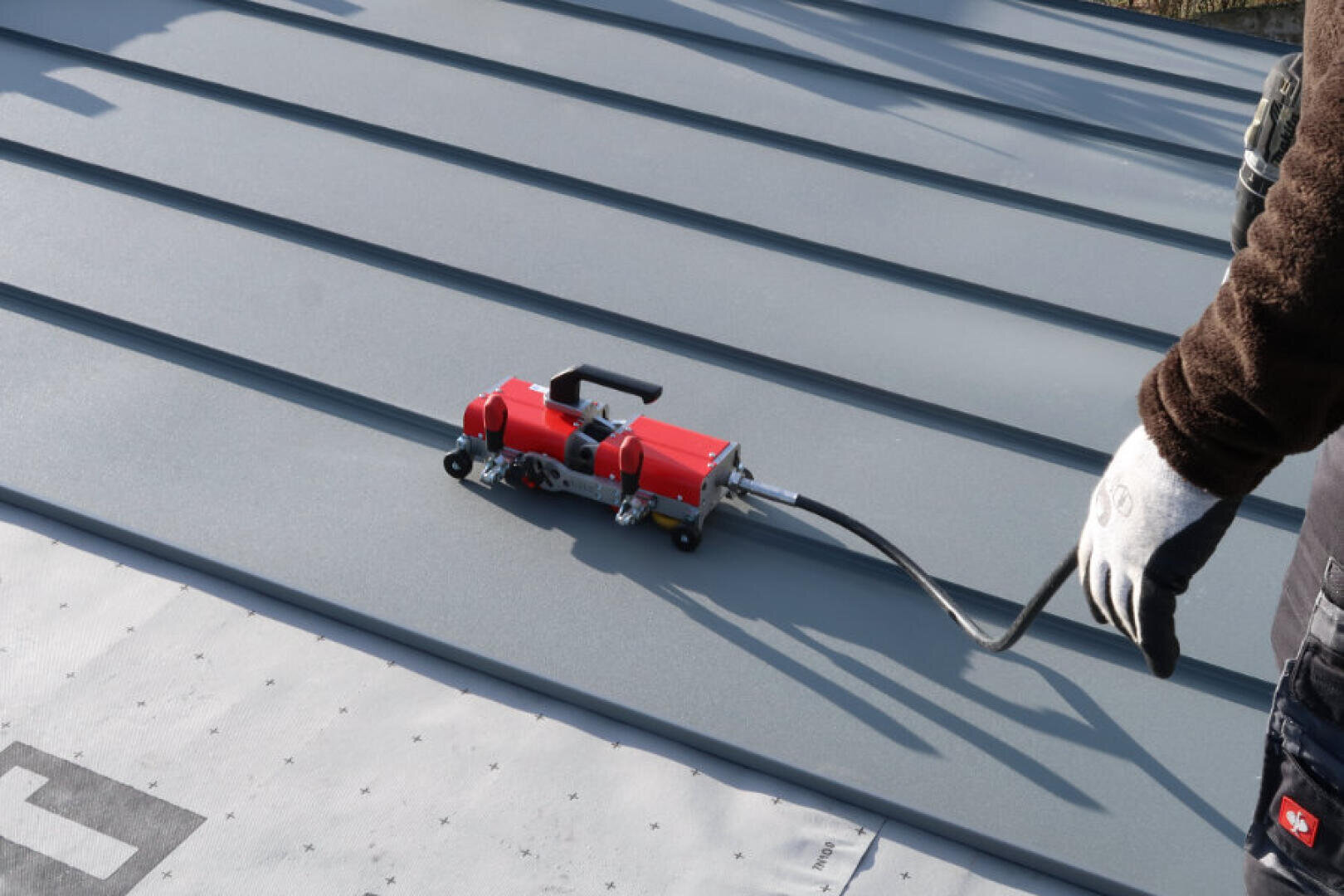 Eine Person mit Handschuhen bedient einen roten Falzschließer auf einem grauen Metalldach mit Stehfalz. Die Maschine ist mit einer flexiblen Welle verbunden, und im Vordergrund ist Dachmaterial zu sehen.