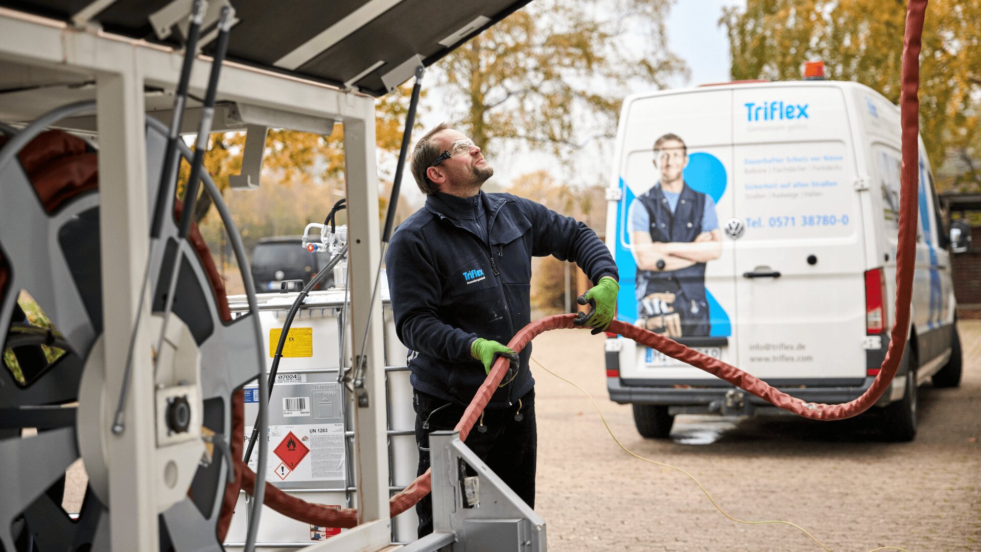Ein Arbeiter in Sicherheitsausrüstung hantiert im Freien mit einem großen roten Schlauch, der an eine Industrieanlage angeschlossen ist. Im Hintergrund sind ein Firmenwagen von Triflex und Herbstbäume zu sehen.