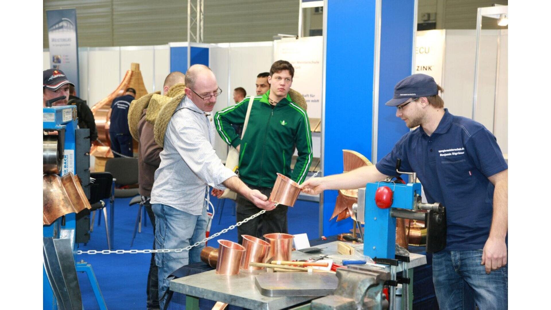 Several men stand around a table with copper items and tools at an exhibition. One man hands a copper vessel to another, as others observe. Exhibits and display boards are visible in the background.