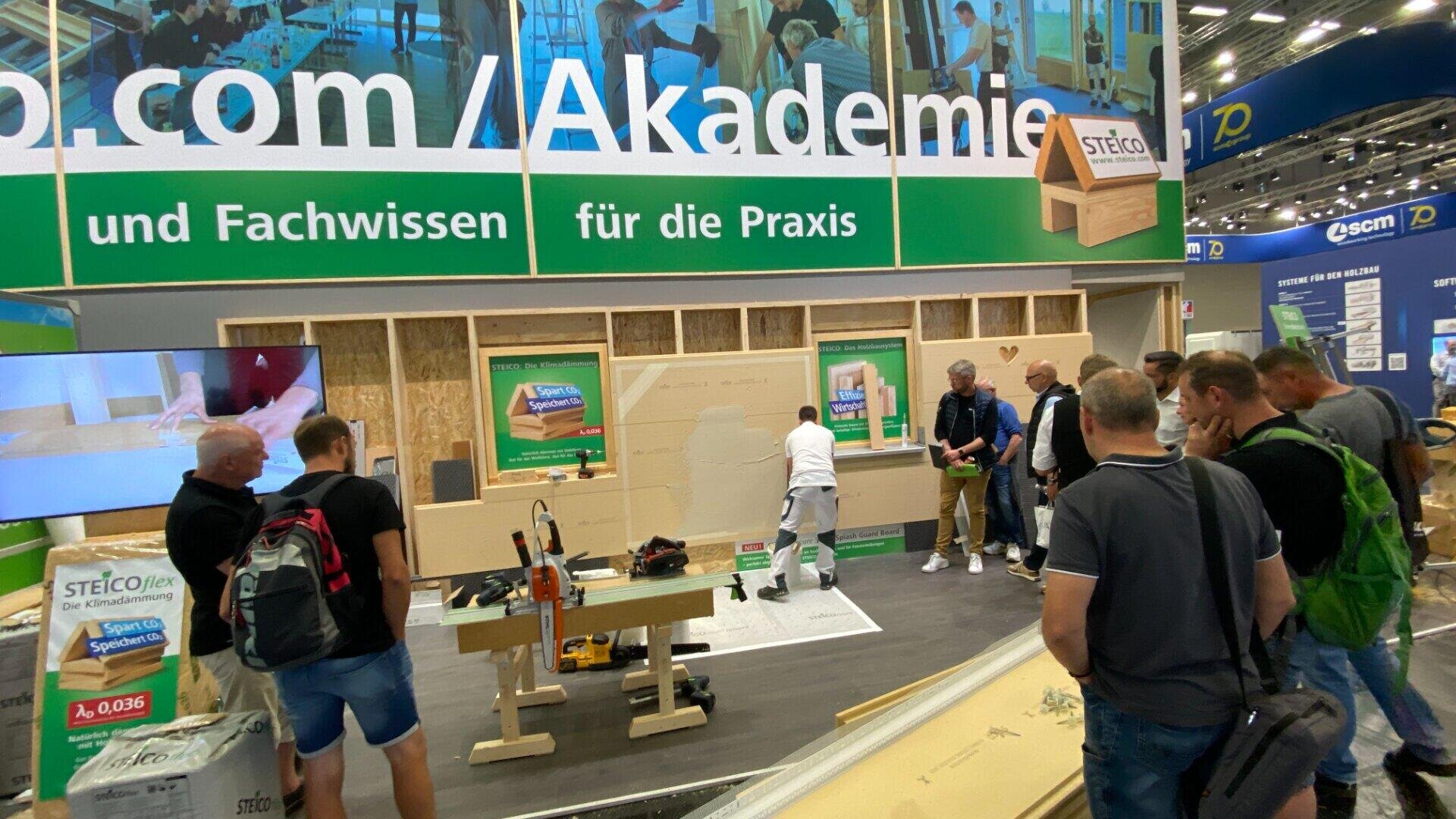 Eine Gruppe von Personen beobachtet eine Live-Vorführung von Bautechniken an einem Messestand mit Werkzeugen, Baumaterialien und Informationsdisplays mit deutschen Texten.